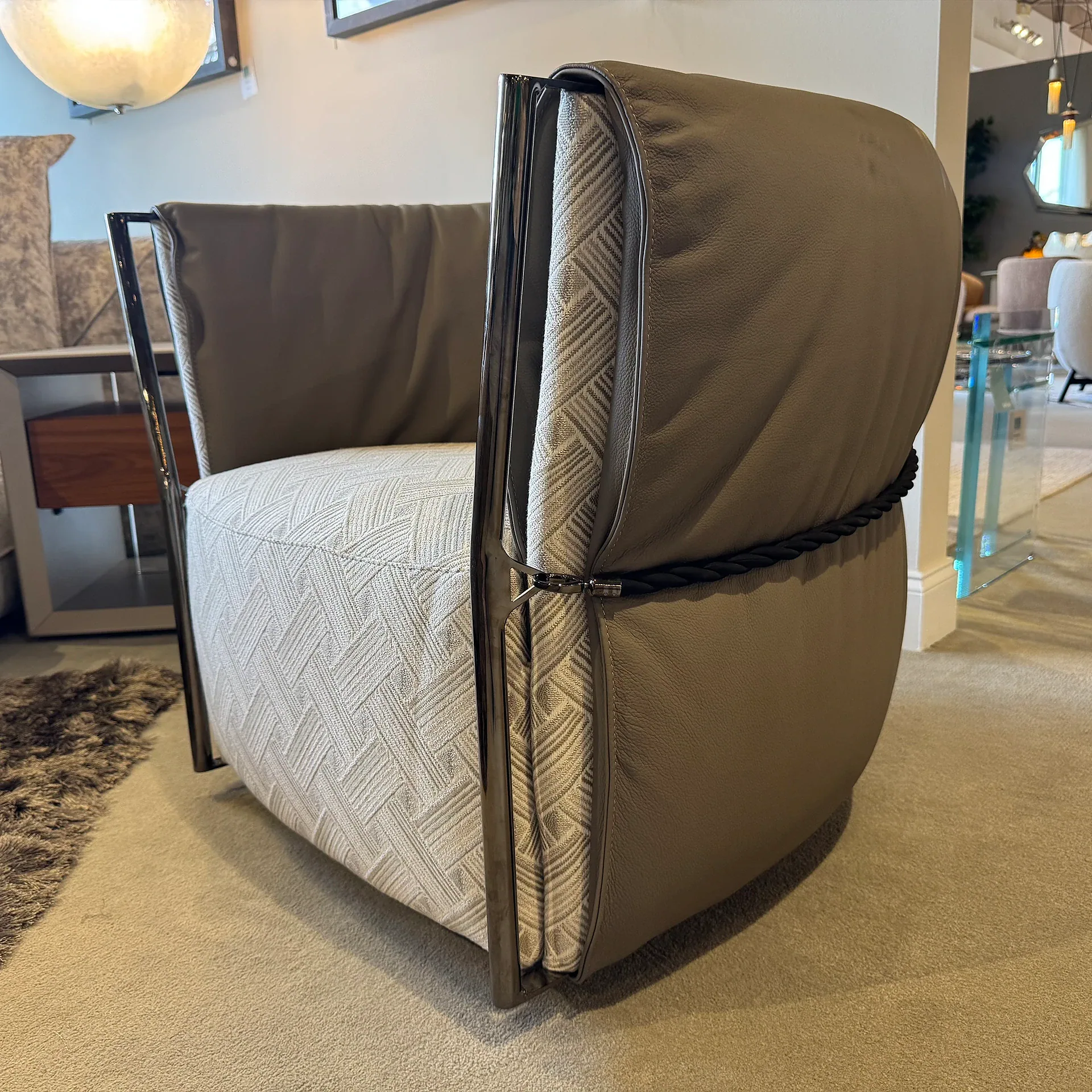 A modern armchair with taupe leather exterior, light patterned fabric interior, dark metal frame, and a black braided rope detail on the back, sitting on a beige carpet.