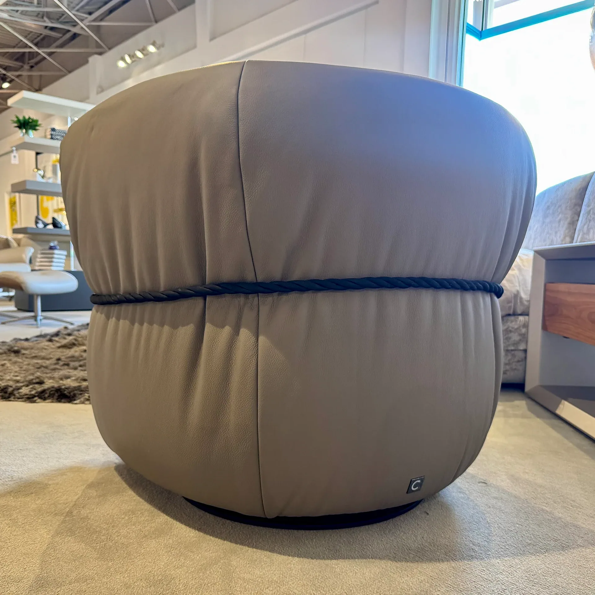 A round, light taupe leather ottoman with a dark braided rope detail around its middle, in a furniture showroom.