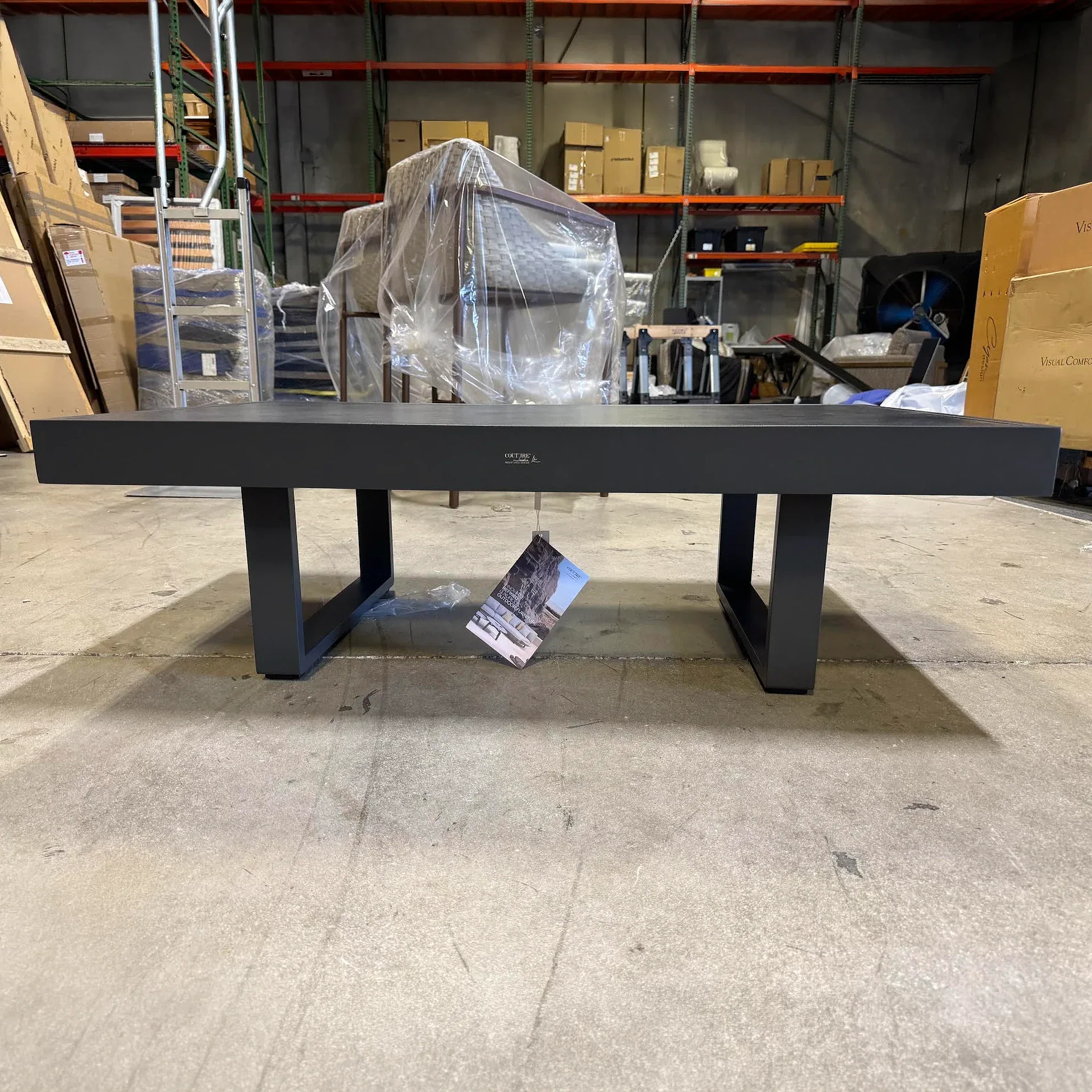 A long, dark gray rectangular coffee table with U-shaped legs sits on a concrete floor in a warehouse, with shelving and wrapped furniture in the background.