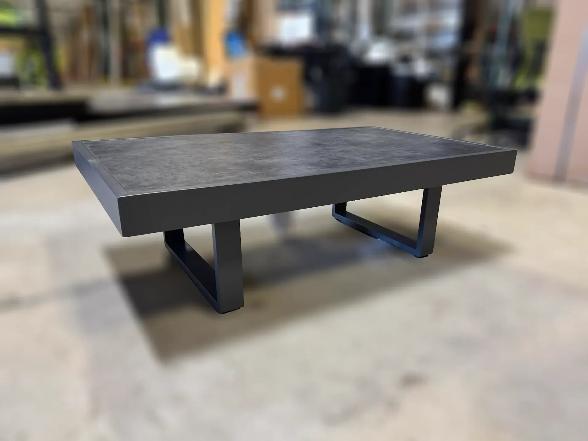 A rectangular coffee table with a dark gray, textured top and matte black U-shaped legs on a light concrete floor.