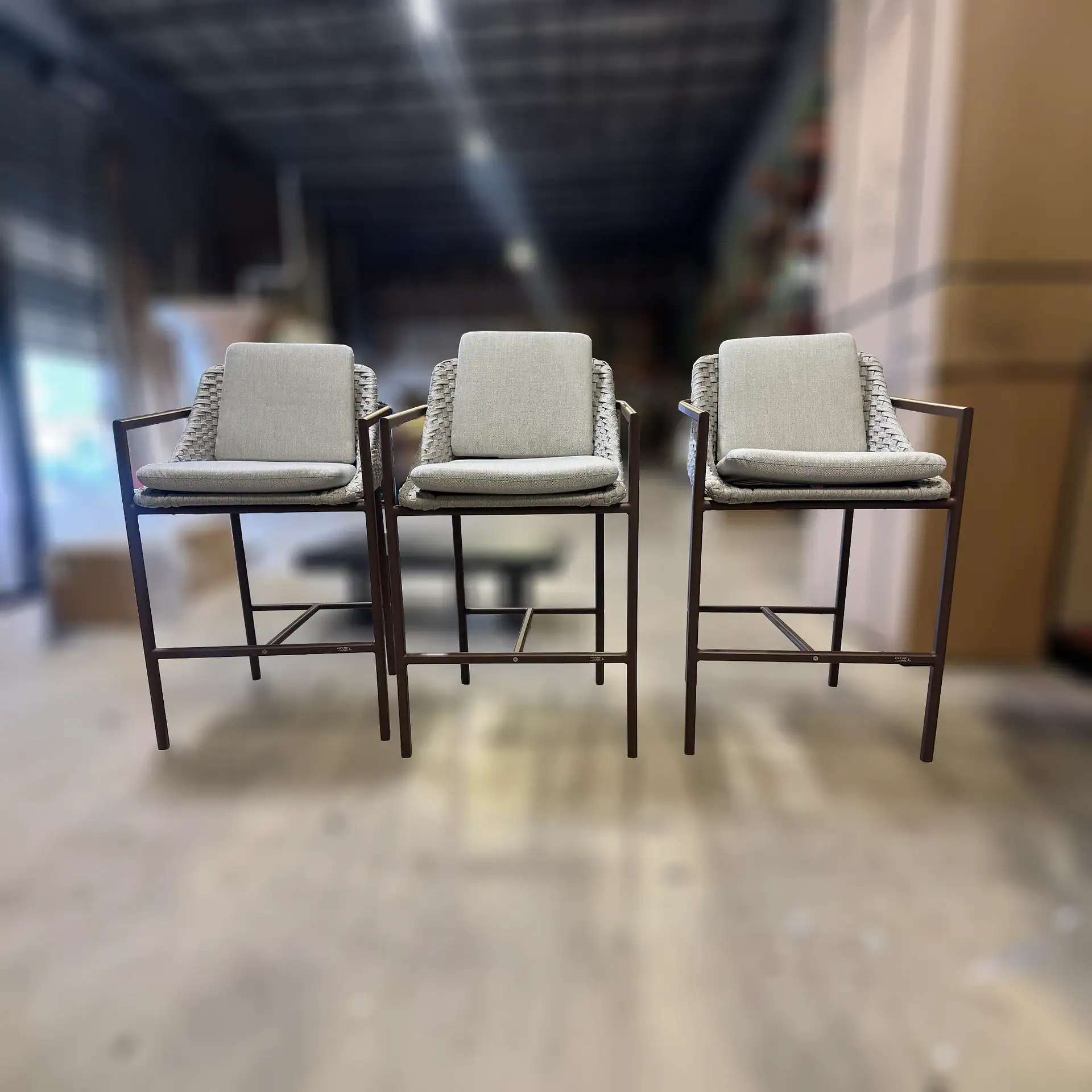 Three light gray bar stools with woven backs and dark metal frames, arranged in a row against a blurred warehouse background.