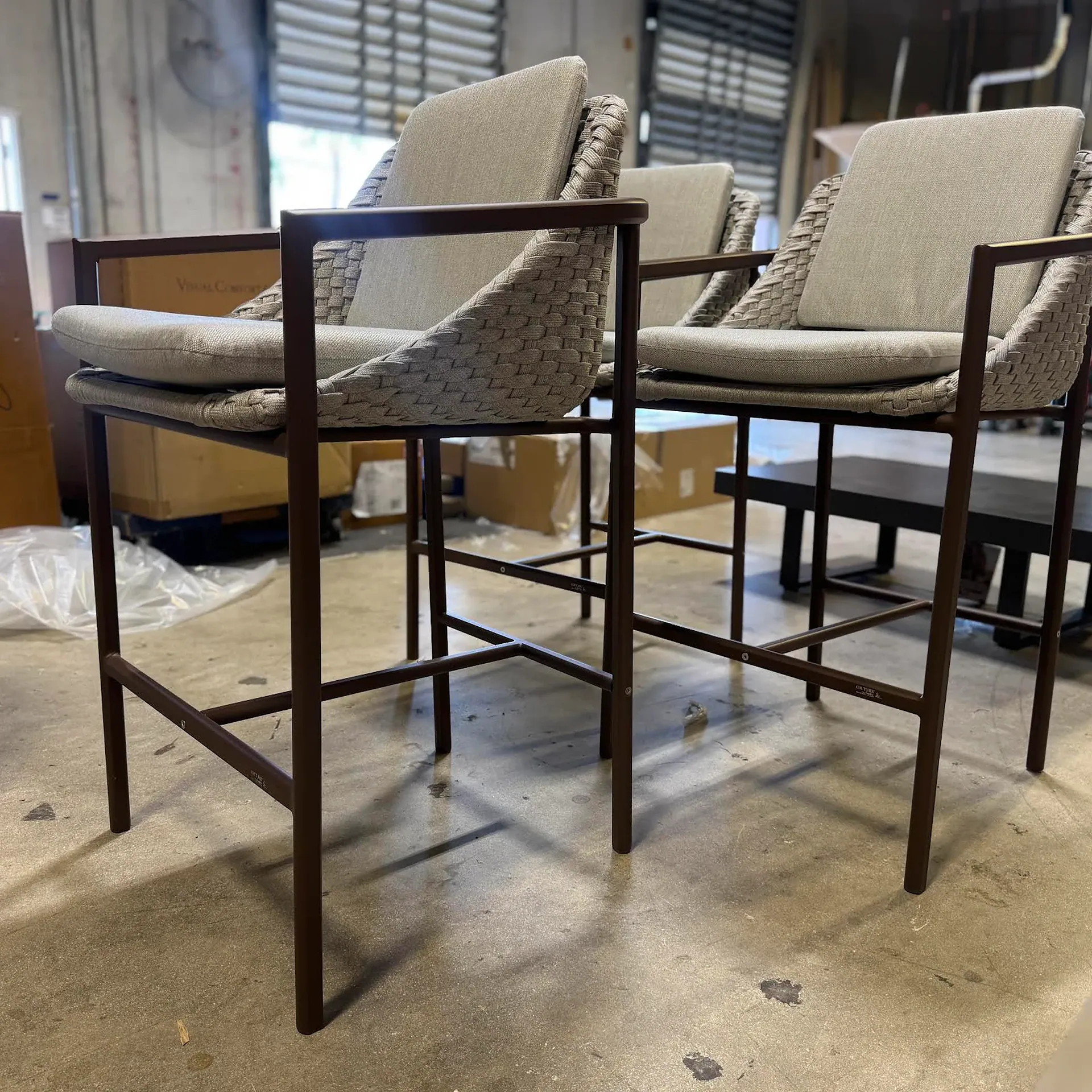 Three modern bar stools with dark metal frames, woven light gray seats, and light fabric cushions on a concrete floor.