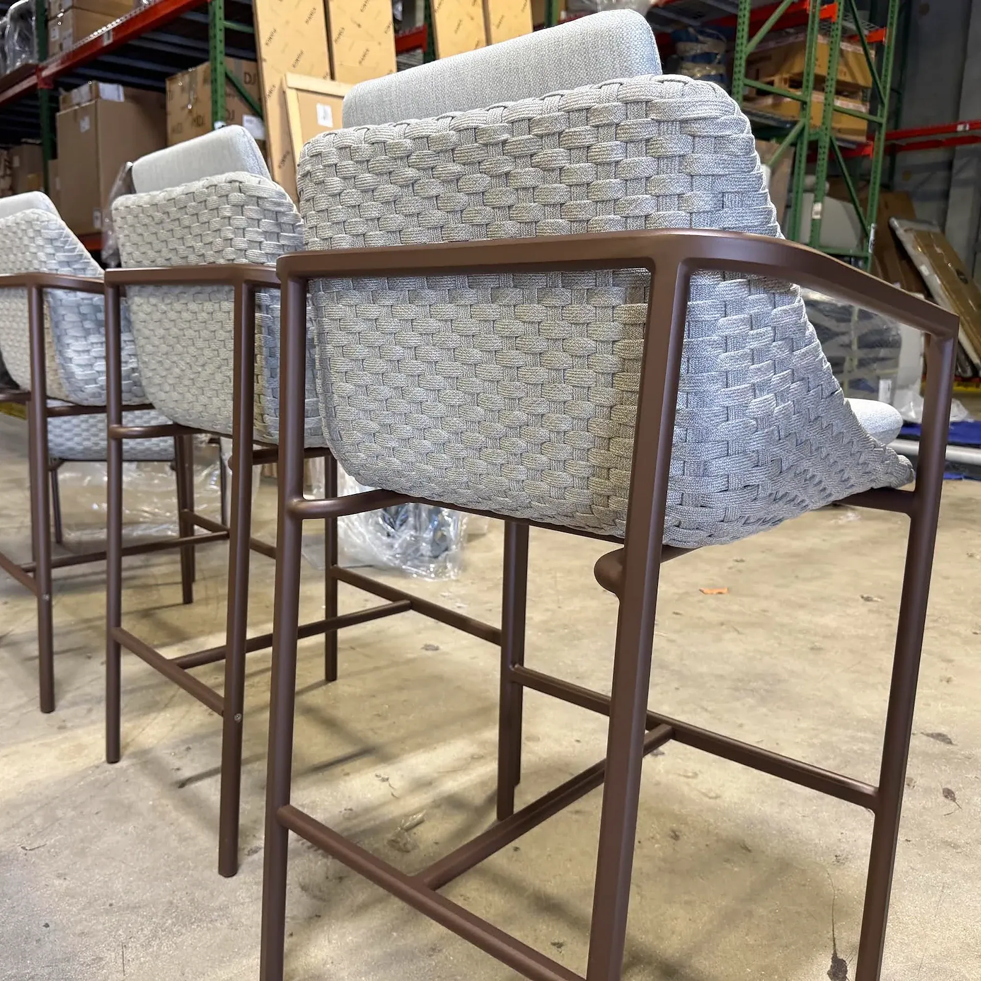 Three modern bar stools with brown metal frames and gray woven seats and cushions, set in a warehouse.