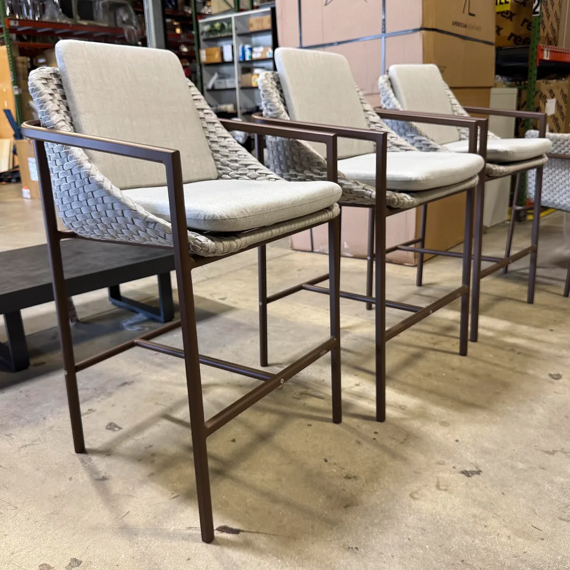 Three contemporary bar stools with dark brown frames, woven light gray backs, and light beige cushions, lined up on a concrete floor in a warehouse.