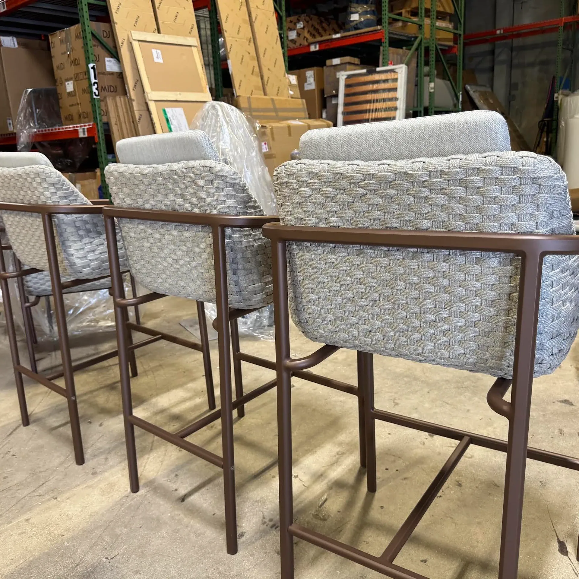 Three light grey woven bar stools with brown frames in a warehouse setting, surrounded by cardboard boxes on shelves.