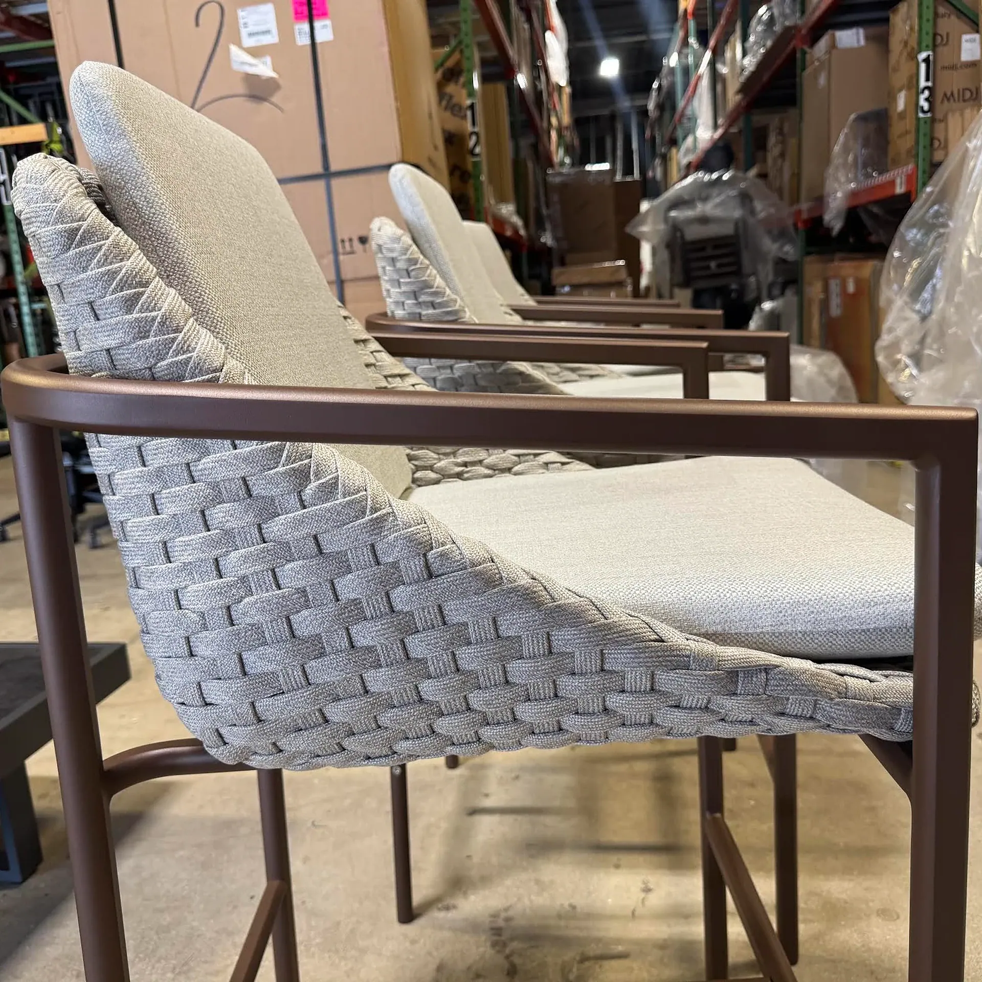 Three modern outdoor bar stools with light gray woven backs and beige fabric seats, featuring dark brown metal frames, lined up in a warehouse.