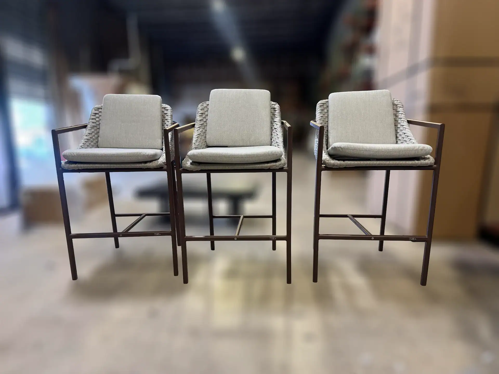 Three identical light grey bar stools with dark metal frames and woven backs, lined up in a row on a light floor.