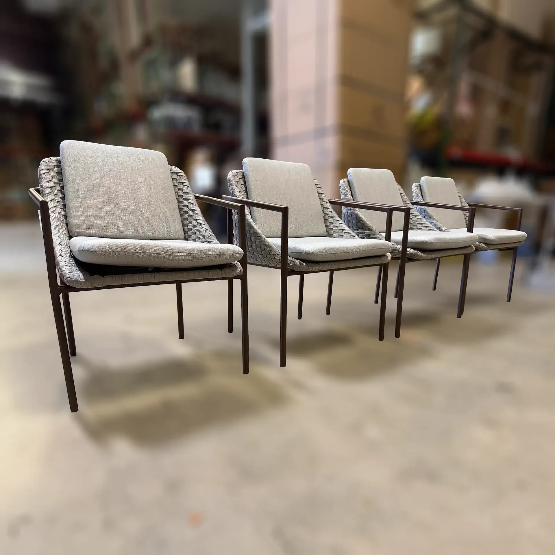 Four modern chairs with dark frames, woven grey backs, and light grey cushions lined up on a light floor in a blurred warehouse setting.