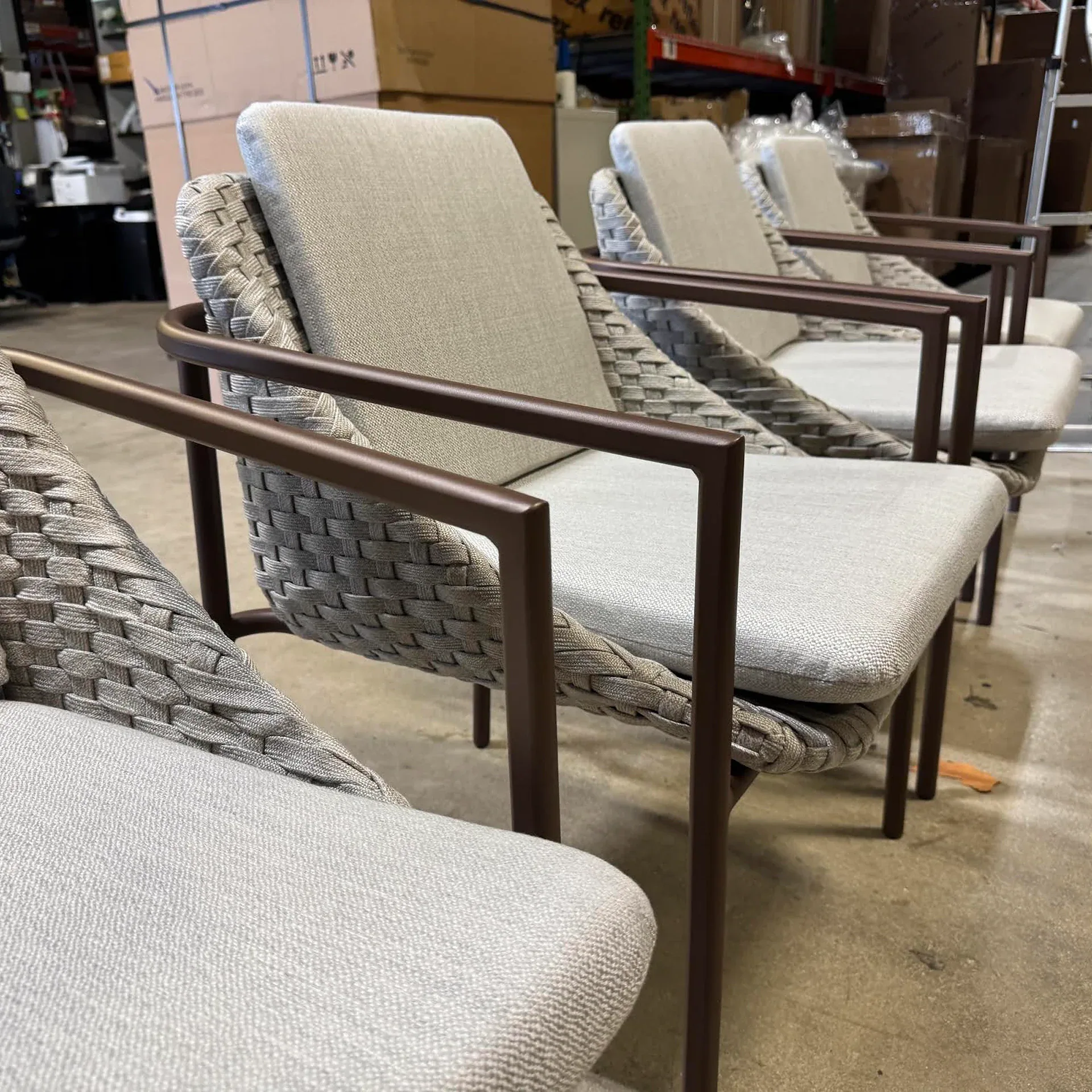 Four modern chairs with dark brown metal frames, light grey woven backs and seats, and light grey cushions, lined up in a warehouse setting.