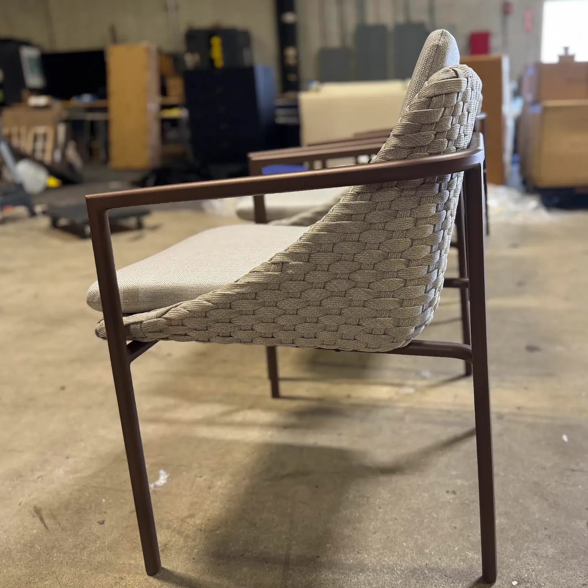 A modern outdoor chair with a dark brown metal frame and light beige woven back and seat, on a concrete floor in a warehouse.