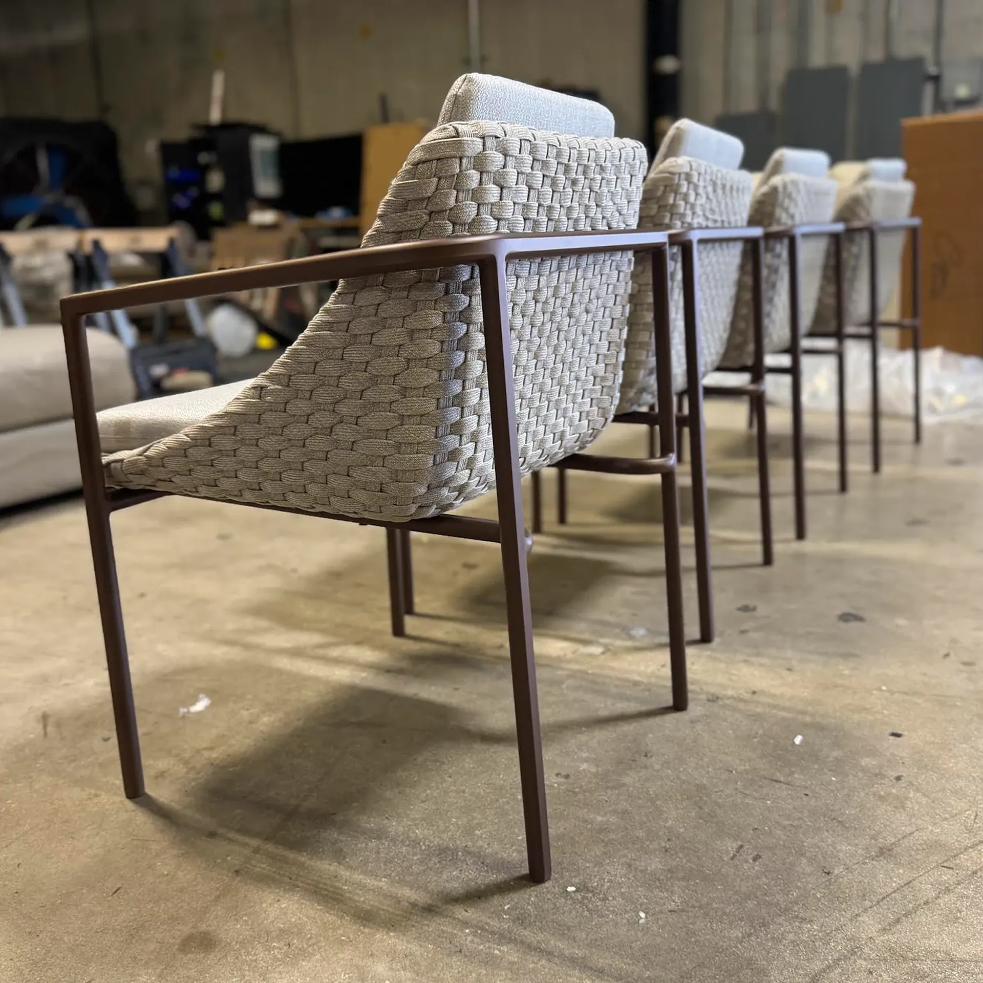 Four modern chairs with woven beige backs, light grey cushions, and dark brown metal frames lined up on a concrete floor.