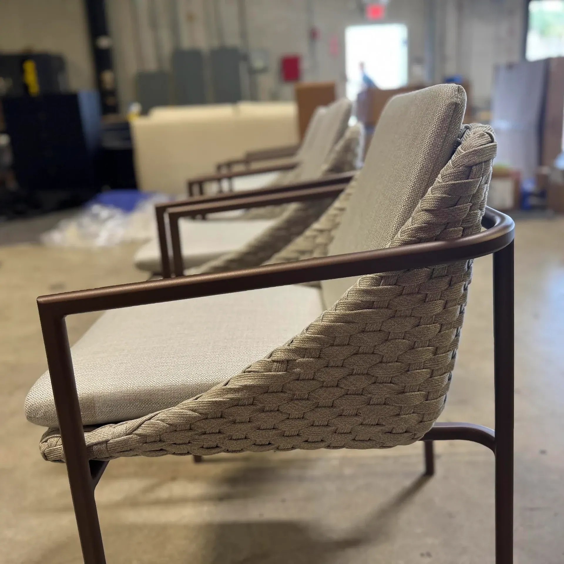 Row of modern armchairs with brown metal frames, woven beige backs, and light grey cushions in a blurred industrial setting.