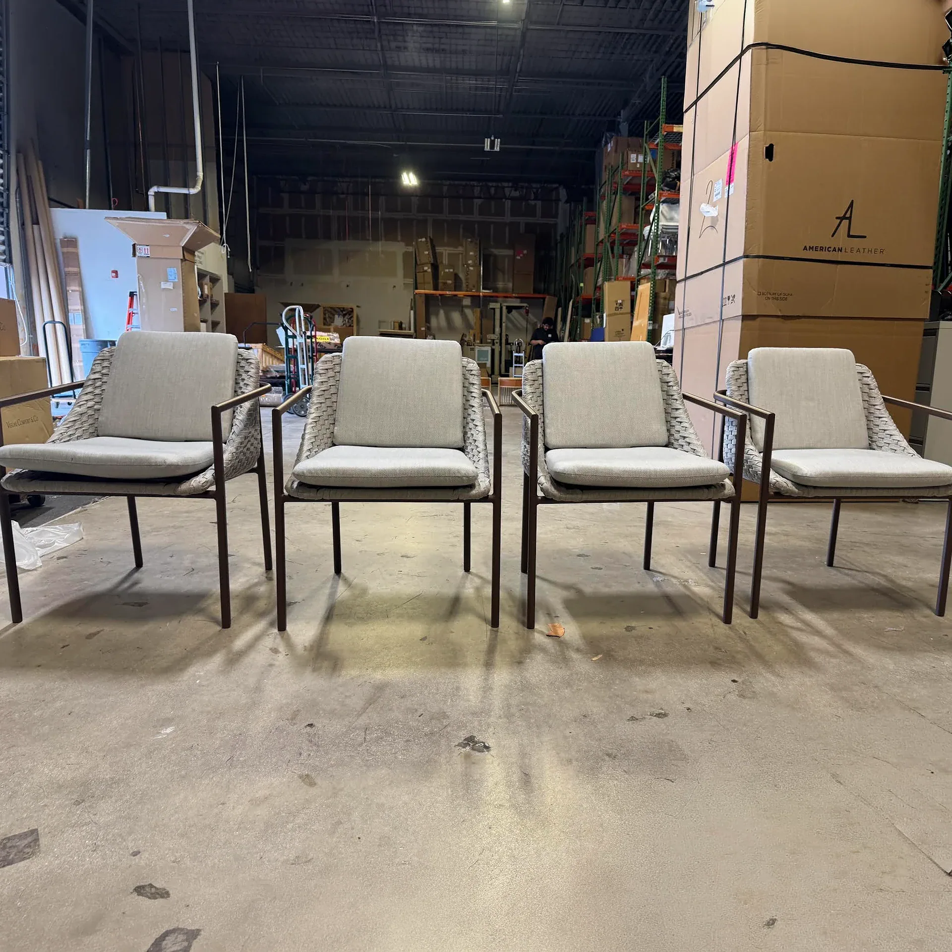 Four modern outdoor armchairs with dark frames and light gray woven backs and cushions, lined up on a concrete floor in a warehouse.