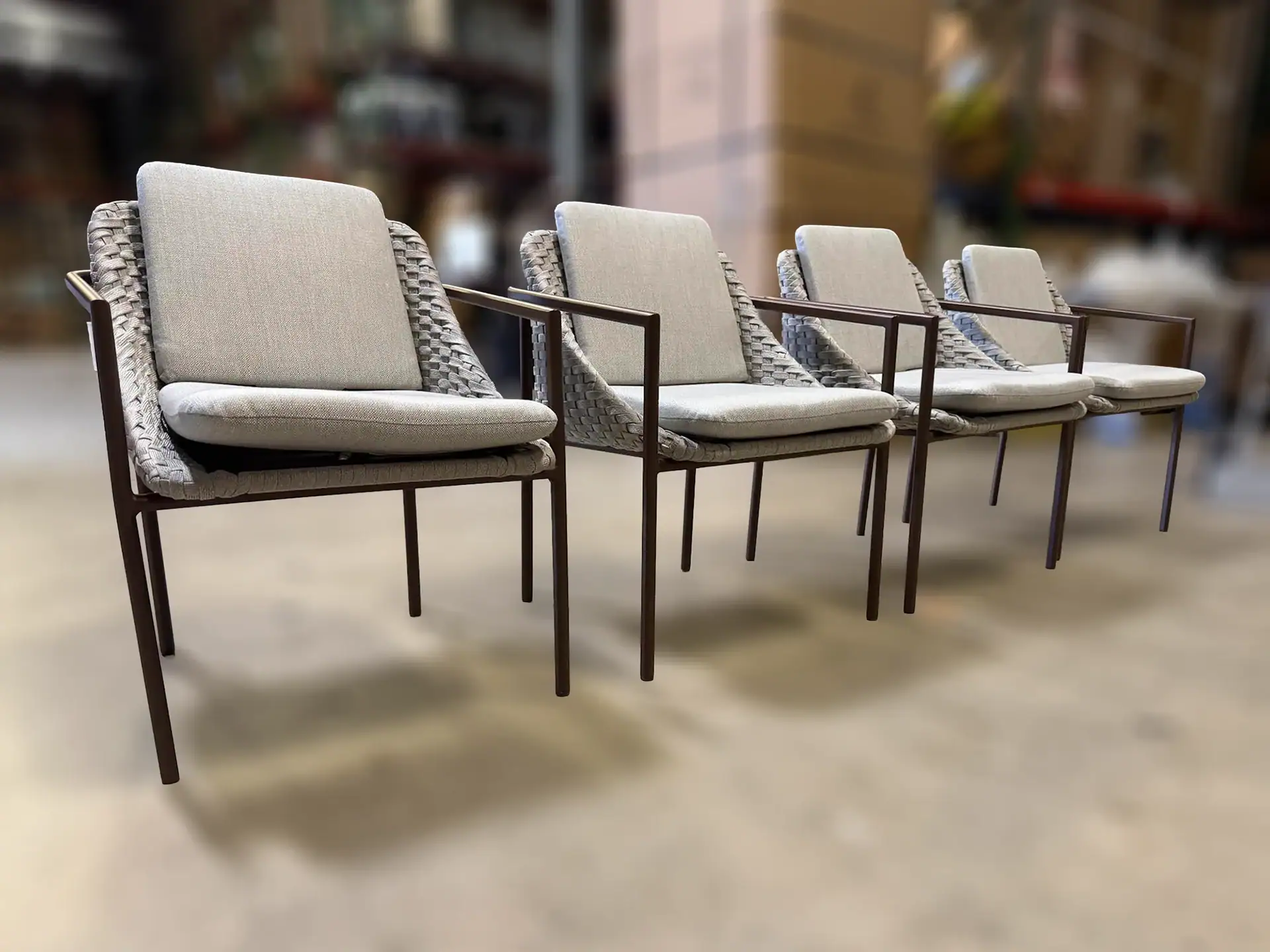 Four identical modern chairs with dark brown frames, woven light gray backs, and light gray cushions, lined up diagonally on a light floor.