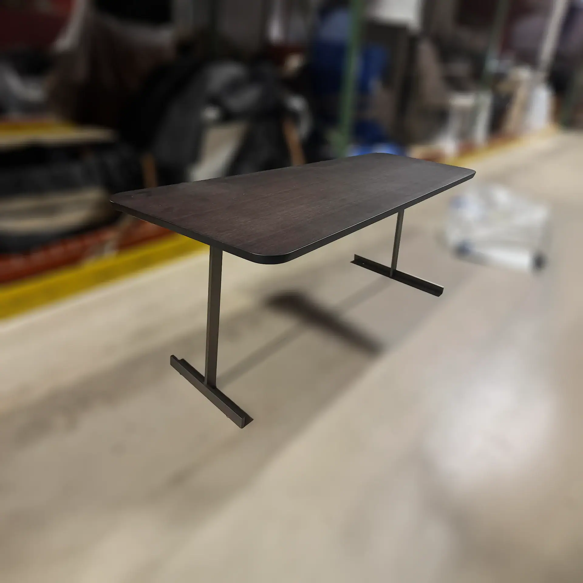 A dark brown rectangular table with rounded corners and two dark metal inverted T-shaped legs, set against a blurred industrial background.