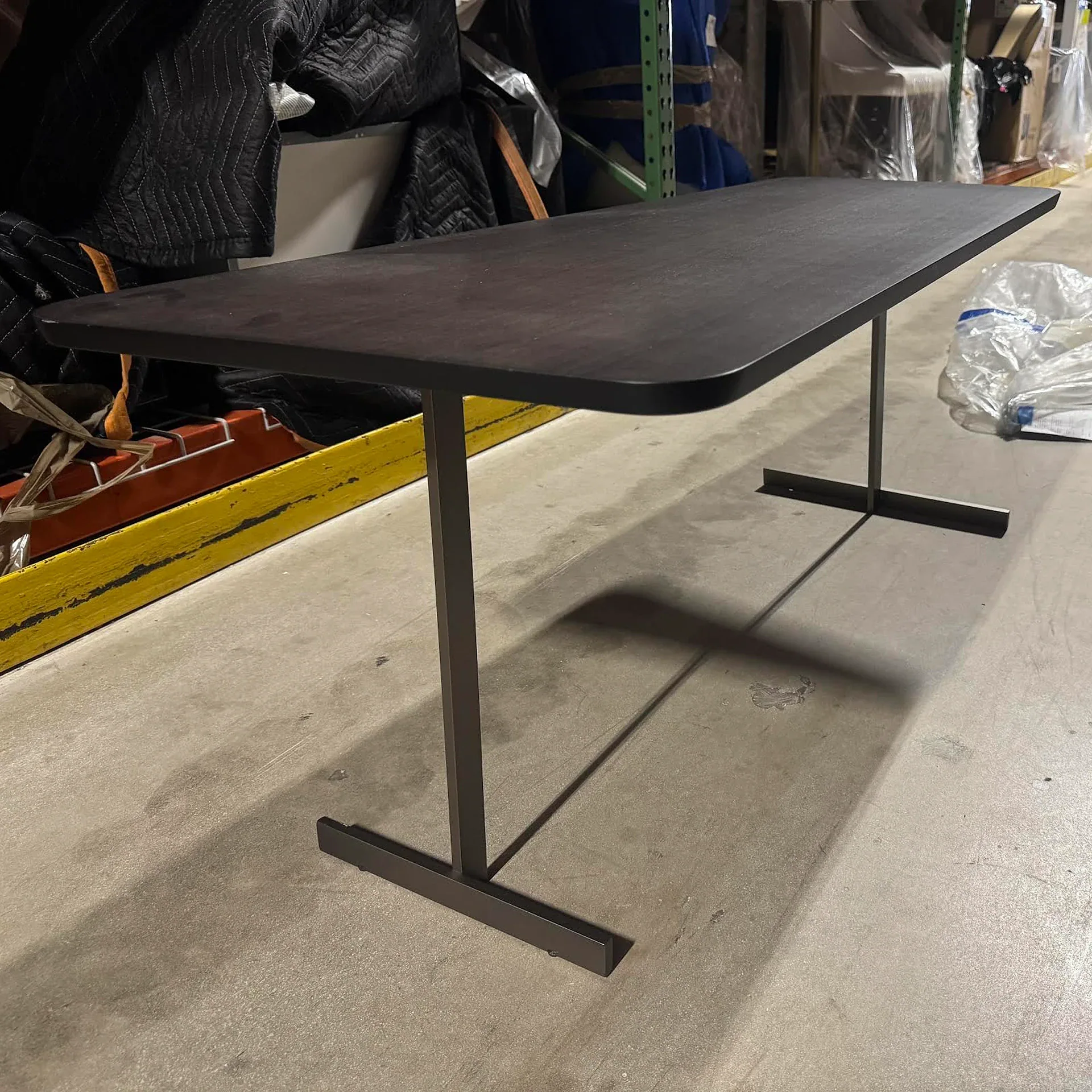 A dark gray rectangular table with rounded corners and T-shaped metal legs stands on a concrete floor in a storage area.