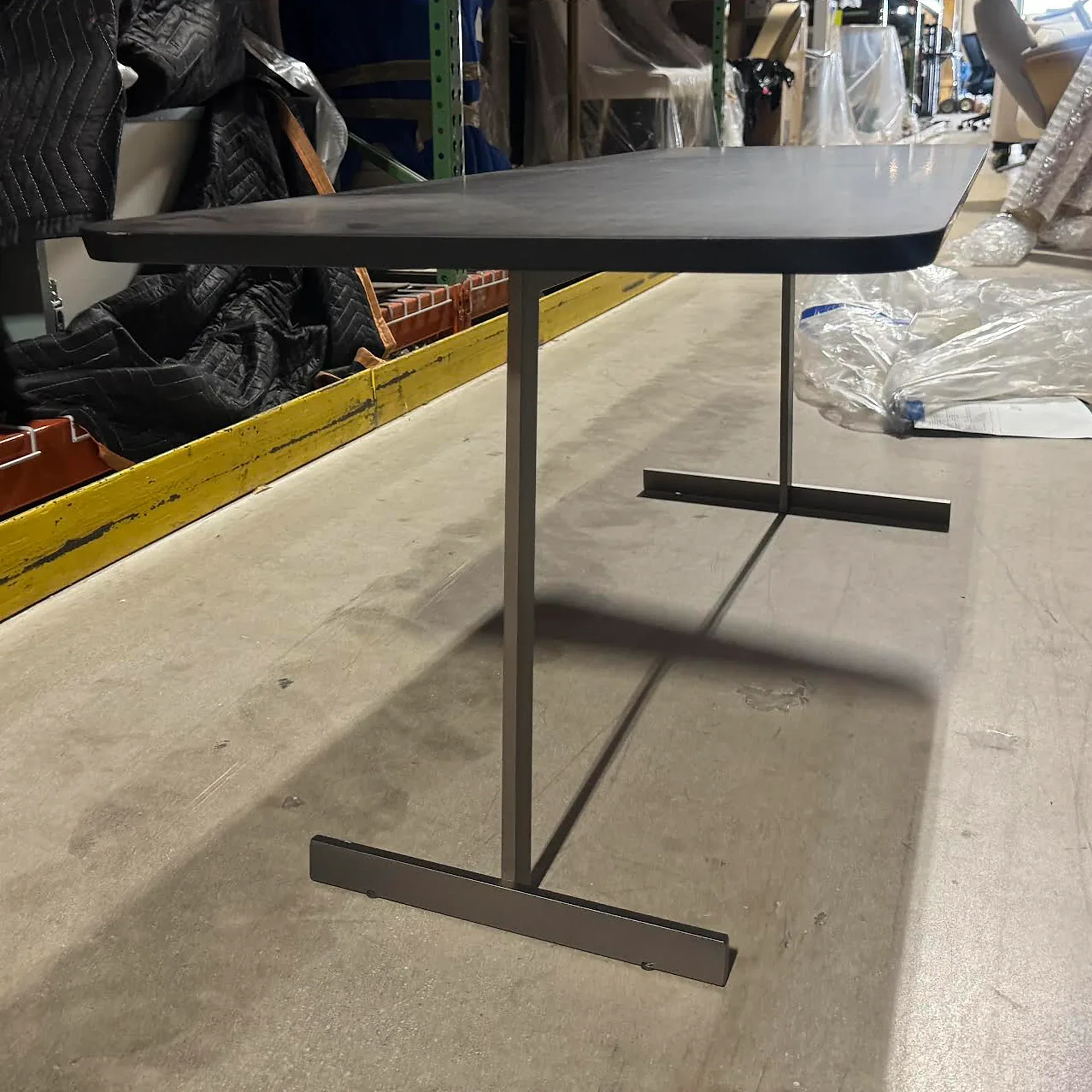 Dark gray rectangular table with metal T-shaped legs on a concrete floor in a warehouse.