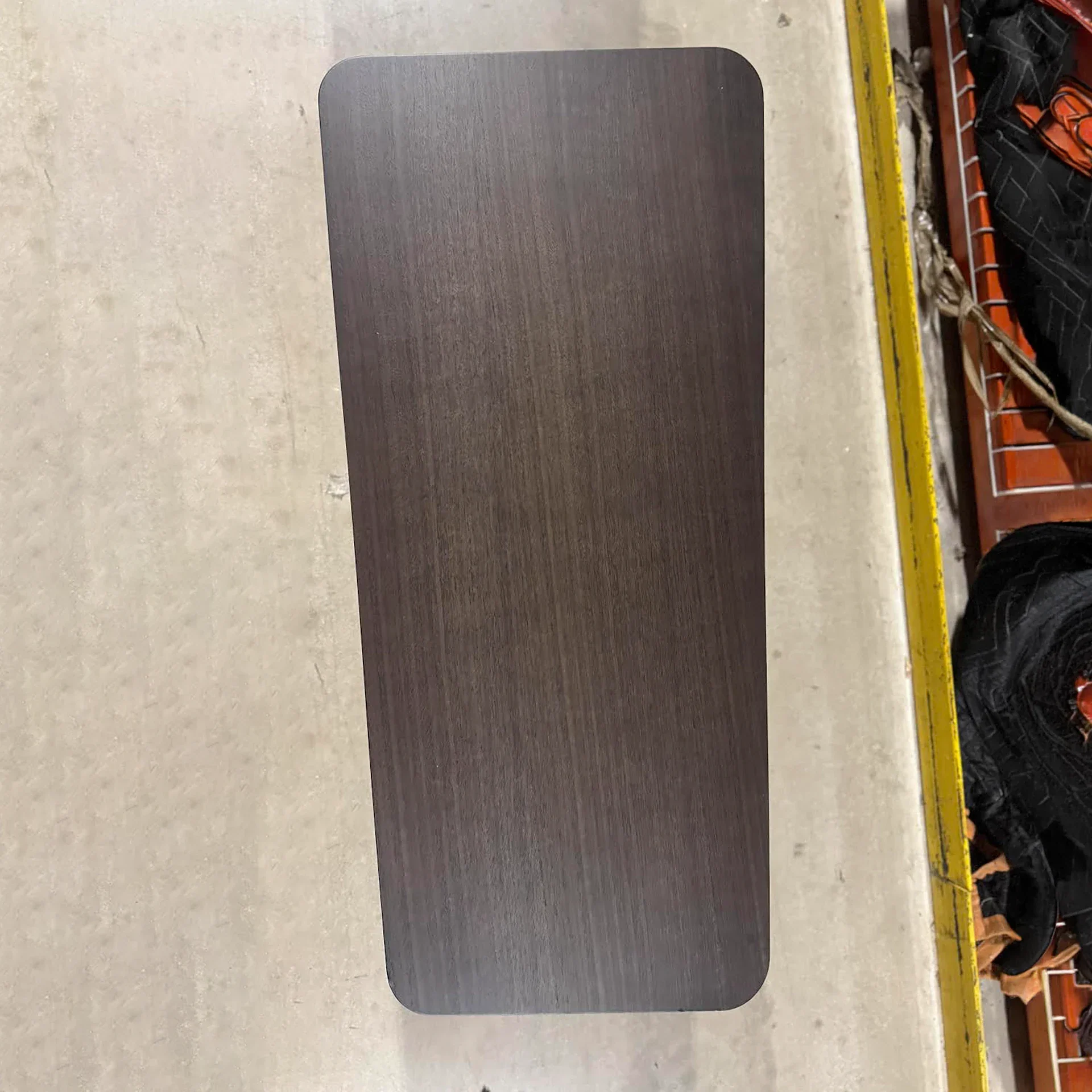 Overhead view of a dark brown rectangular panel with rounded corners on a light concrete floor, next to a yellow beam and cluttered items.