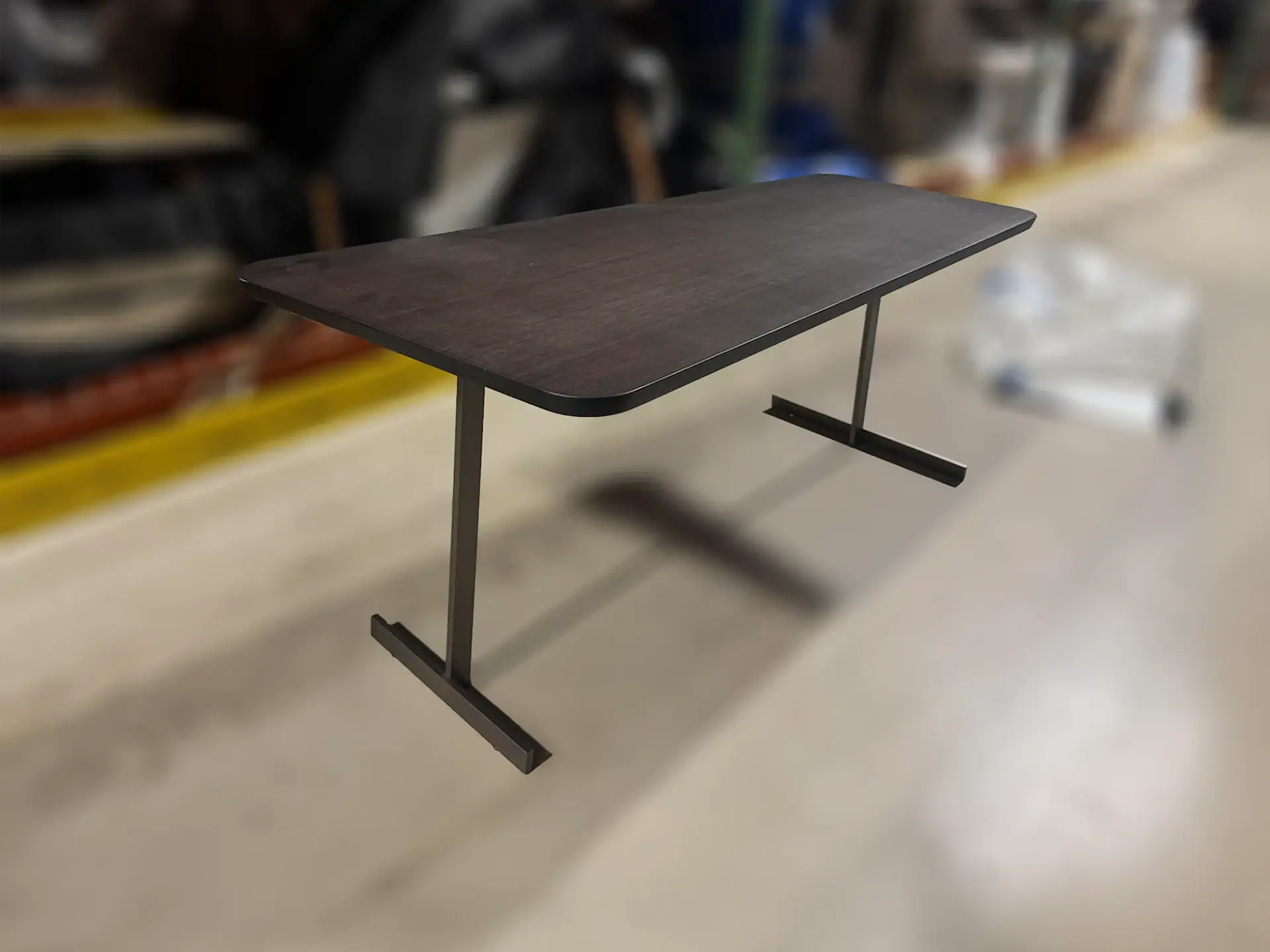 A dark brown rectangular table with rounded corners and two dark metal T-shaped legs on a light-colored floor with a blurred background.