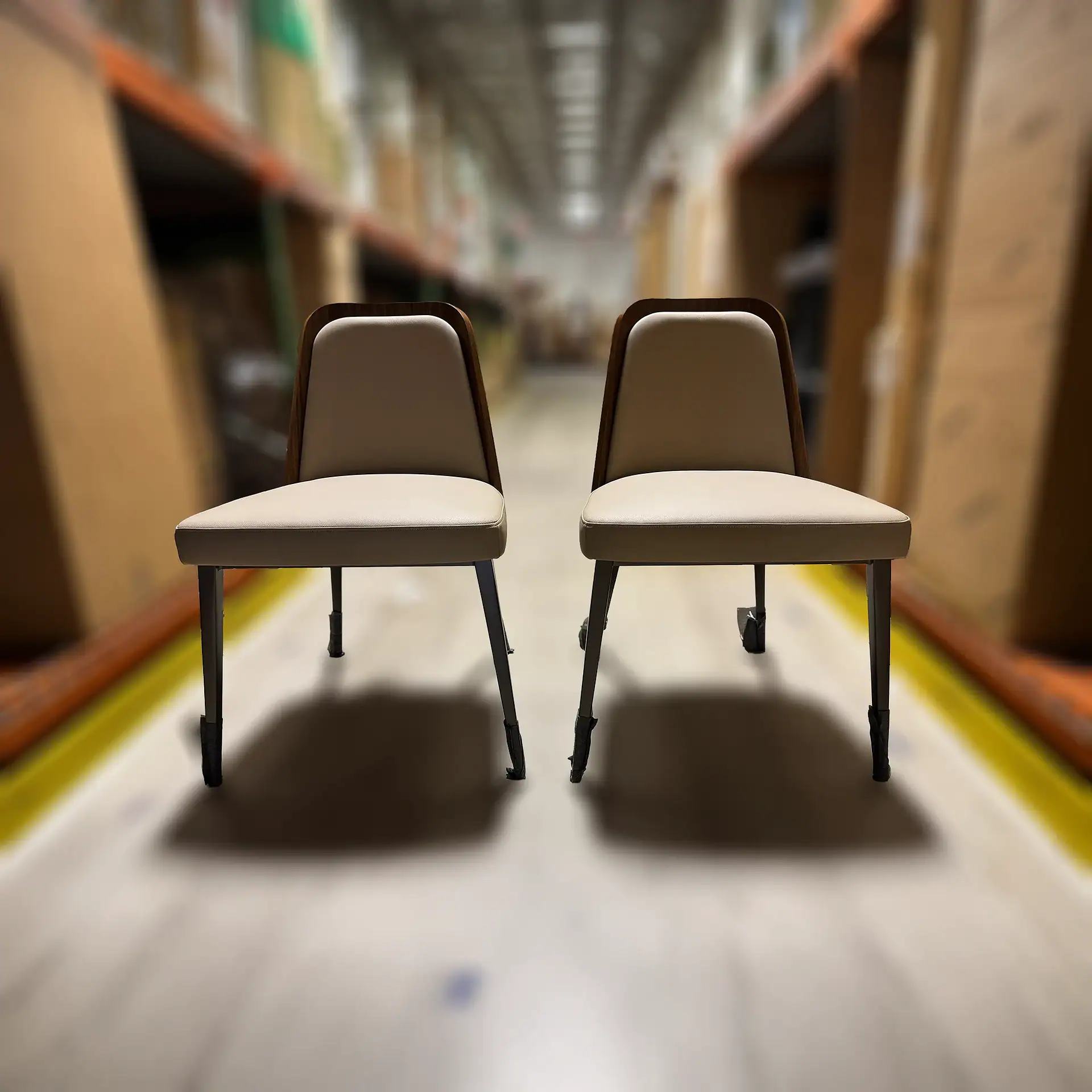 Two beige and dark brown chairs with black legs sit side-by-side in a blurred warehouse aisle with shelves of boxes.
