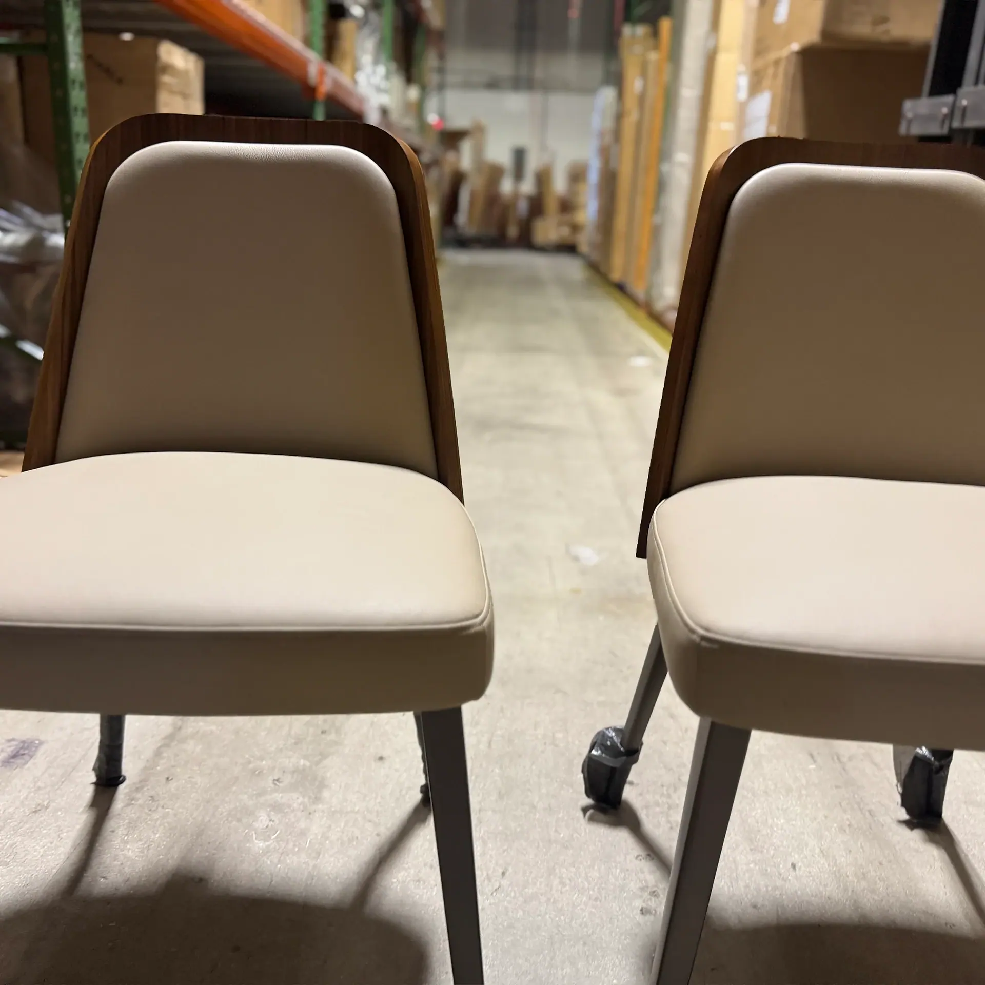 Two modern chairs with beige upholstery and wooden backs on a concrete floor in a warehouse.