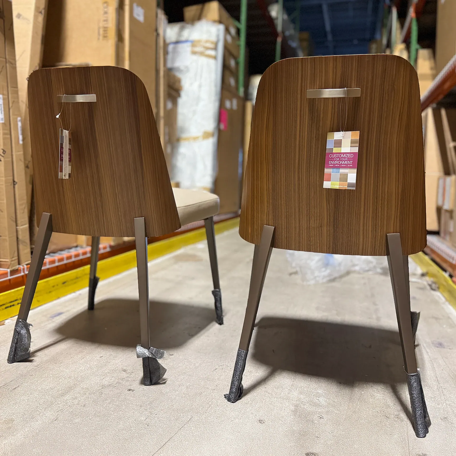Two modern chairs with wood-grain backrests, metal legs, and customization tags, seen from behind in a warehouse.