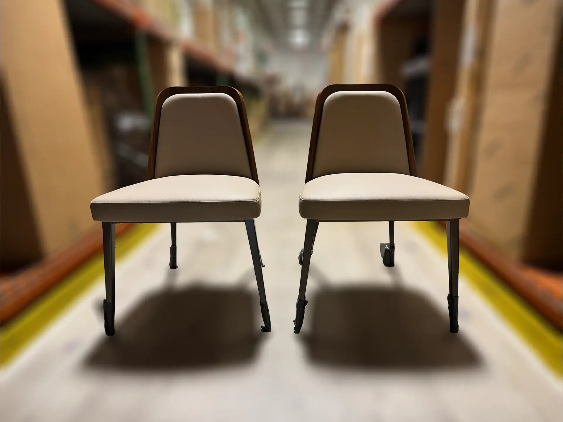 Two light beige upholstered chairs with dark wood backs and metal legs stand side-by-side in a blurred warehouse aisle with shelves of boxes.