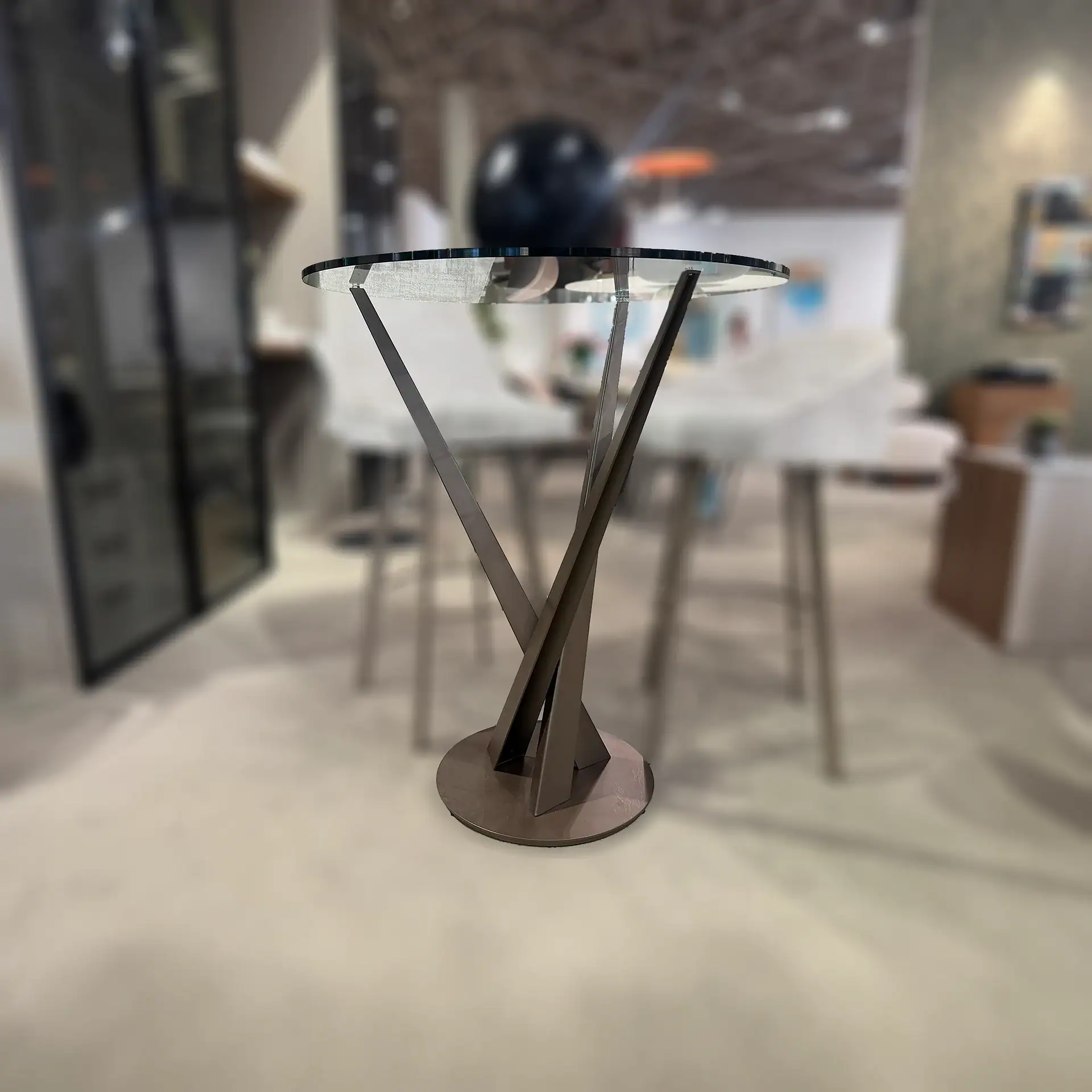A modern round glass-top table with a dark, sculptural metal base twisting upwards from a circular plate. The background is blurred.