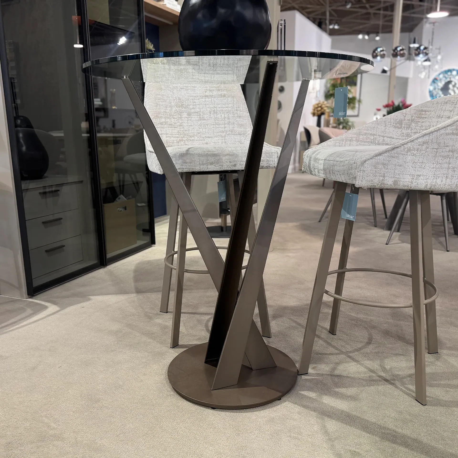 A modern bar-height table with a glass top and unique metallic base, flanked by two upholstered bar stools, in a furniture showroom.