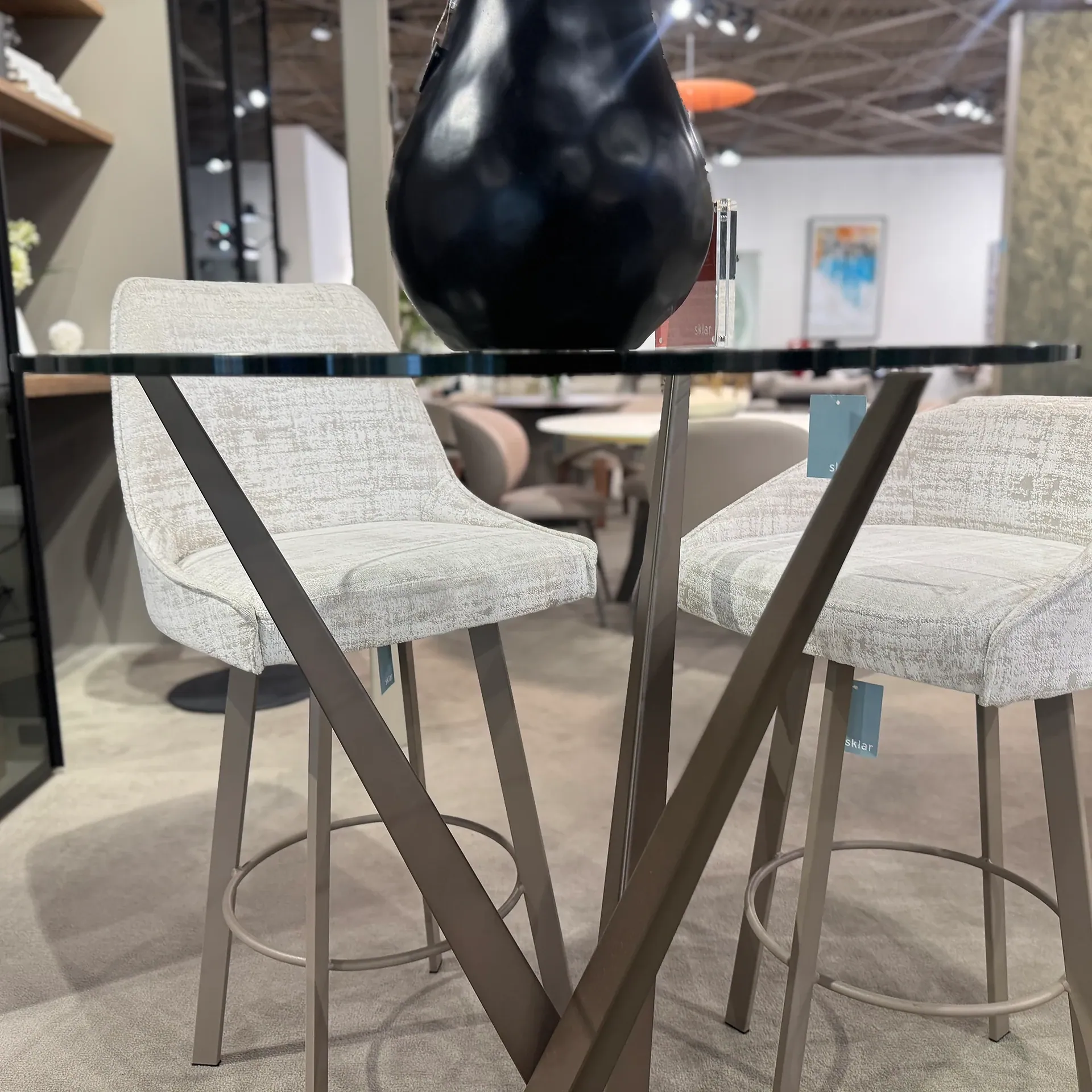 A modern bar table with a glass top and metallic base, flanked by two upholstered bar stools, topped with a black vase in a showroom setting.