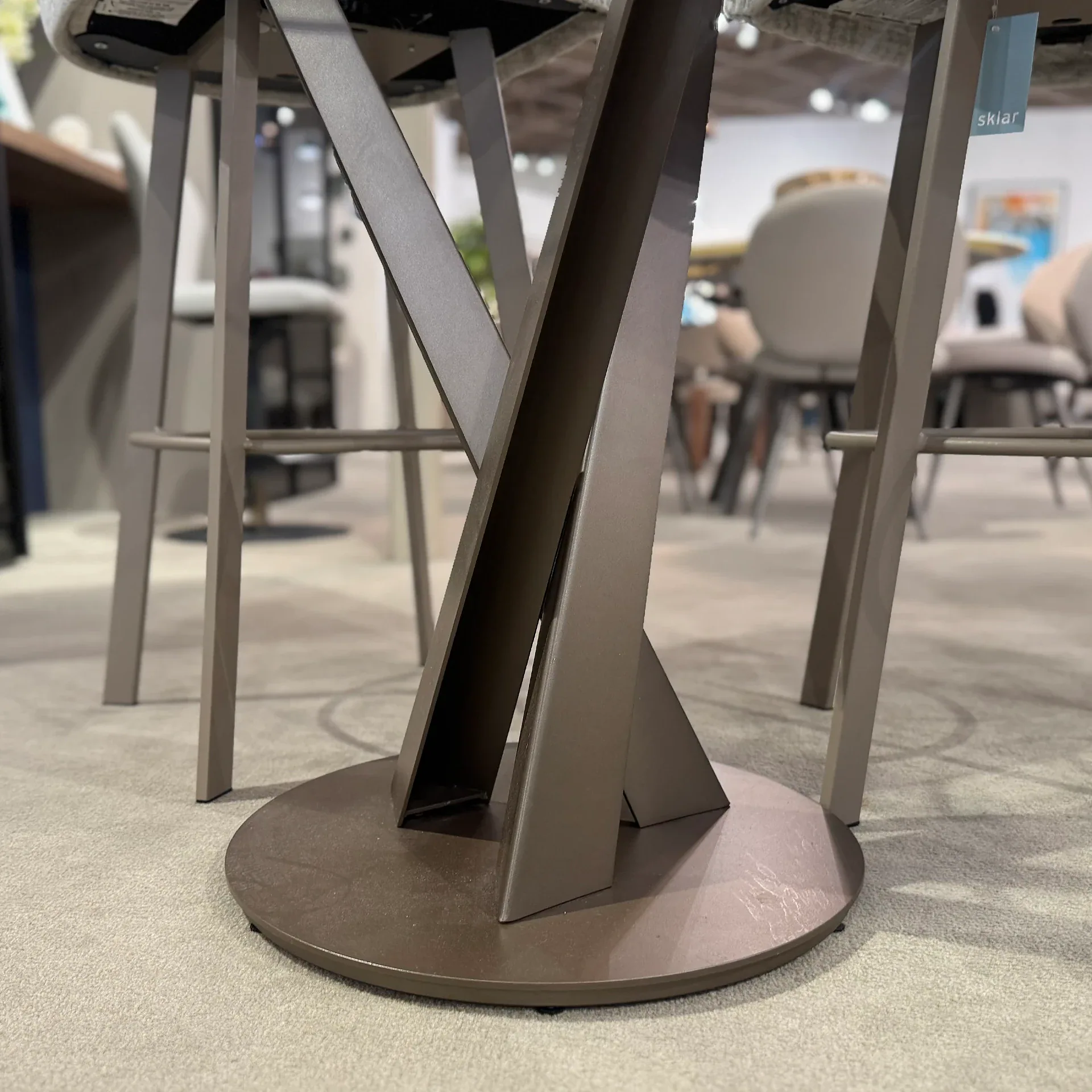 Close-up of a dark bronze or taupe metal furniture base with a round bottom and criss-crossing legs, on a light gray carpet. A tag with 'sklar' hangs from one leg.