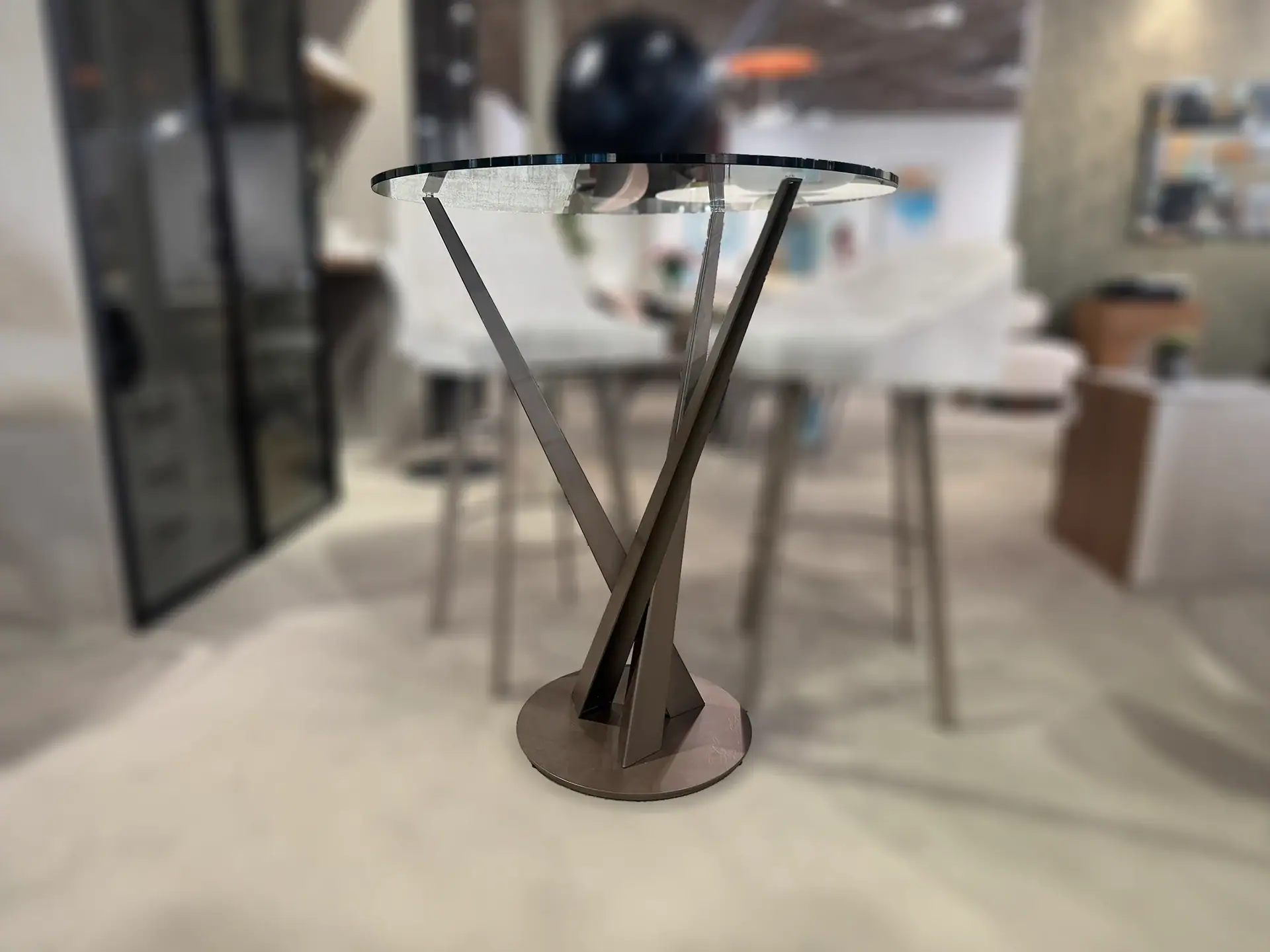 A round glass-top table with a unique dark bronze twisted metal base stands on light flooring in a blurred room.