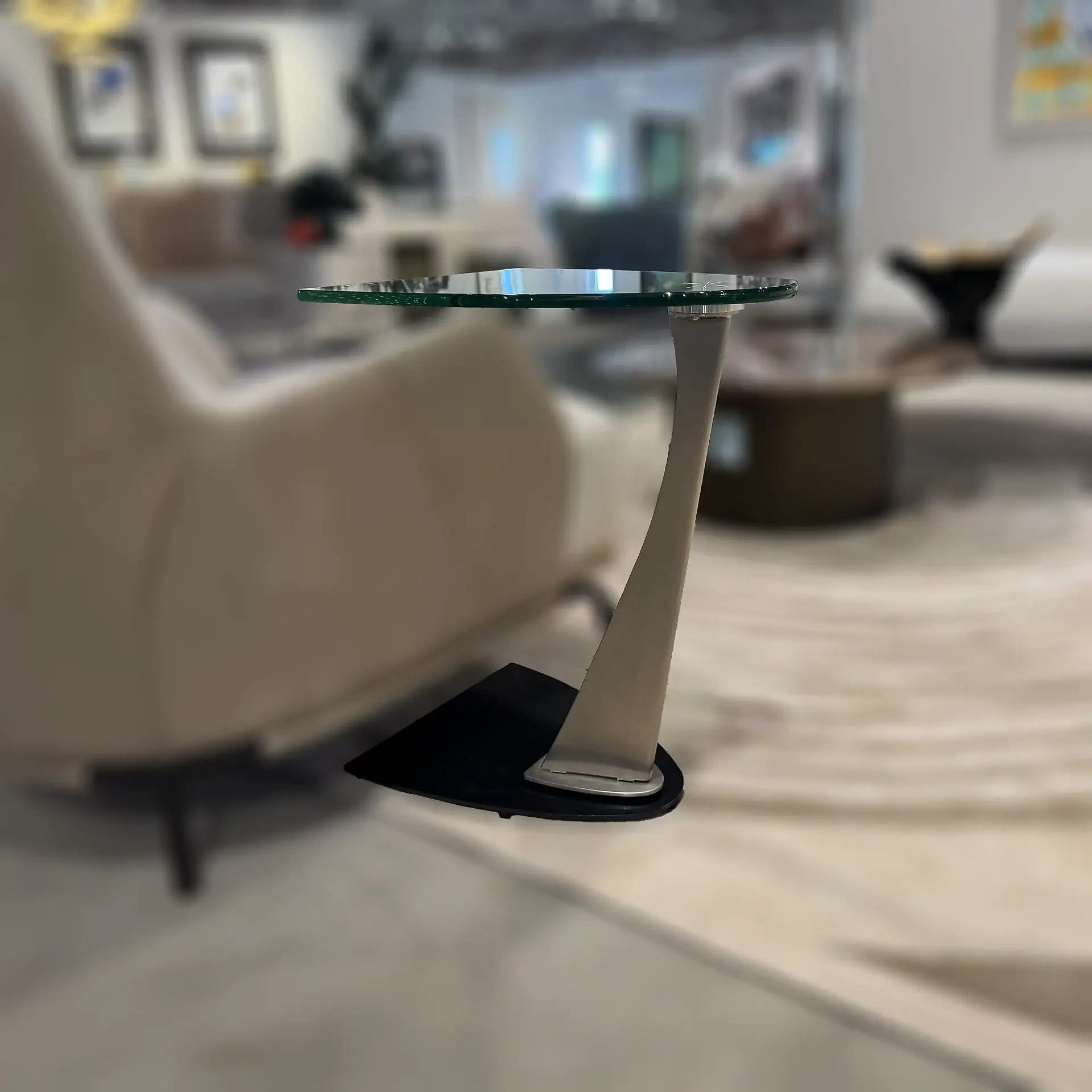 A modern cantilevered side table with a round glass top and a curved, brushed silver-toned metal base on a light rug in a blurred room.