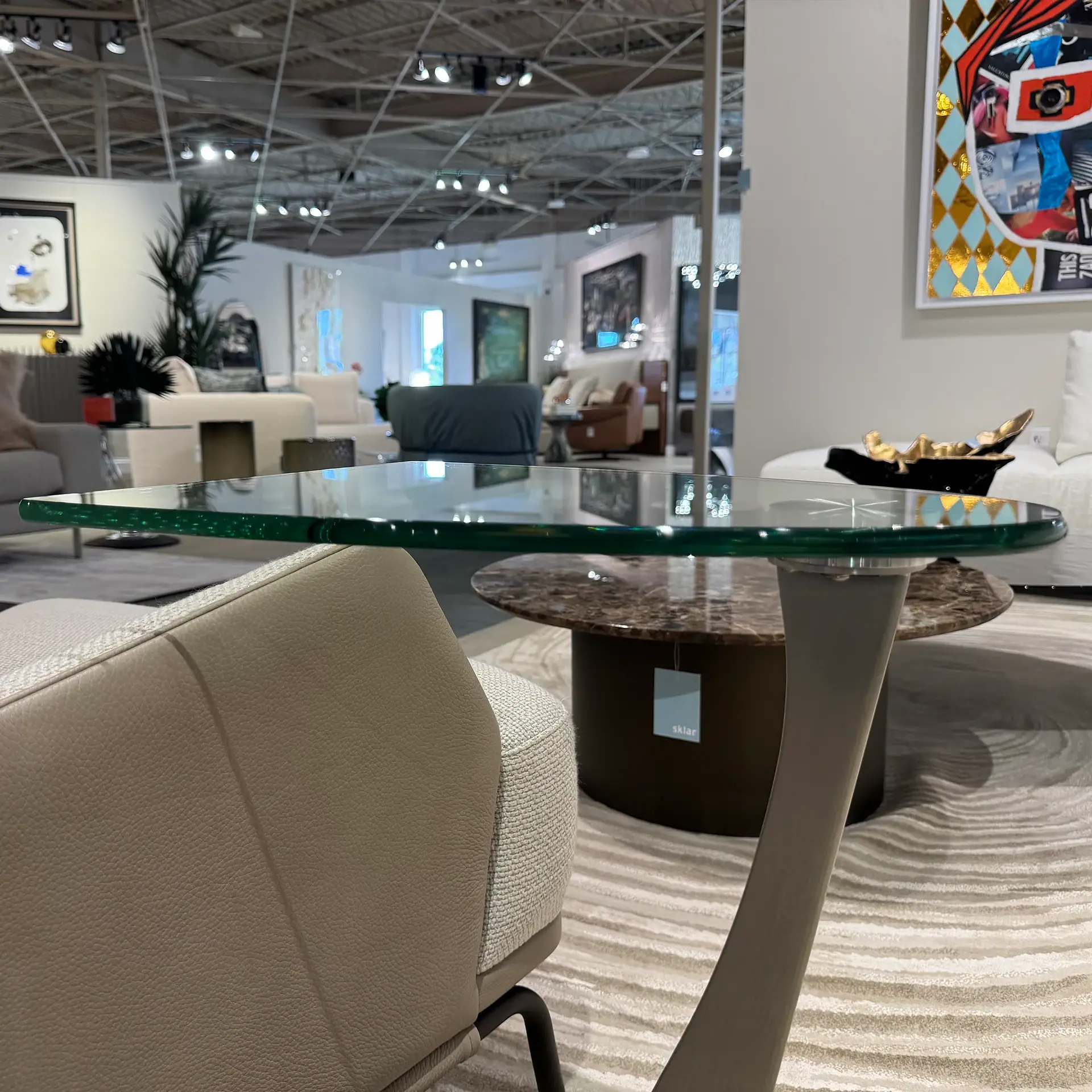 A modern furniture showroom scene featuring a glass-top coffee table, a marble-top table with a 'sklar' tag, and a beige armchair on a wavy patterned rug.