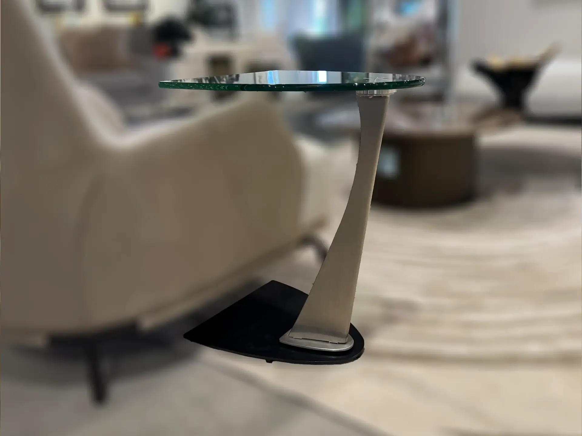 A modern side table with a round glass top, a curved silver metallic stand, and a black base, set against a blurred living room background.