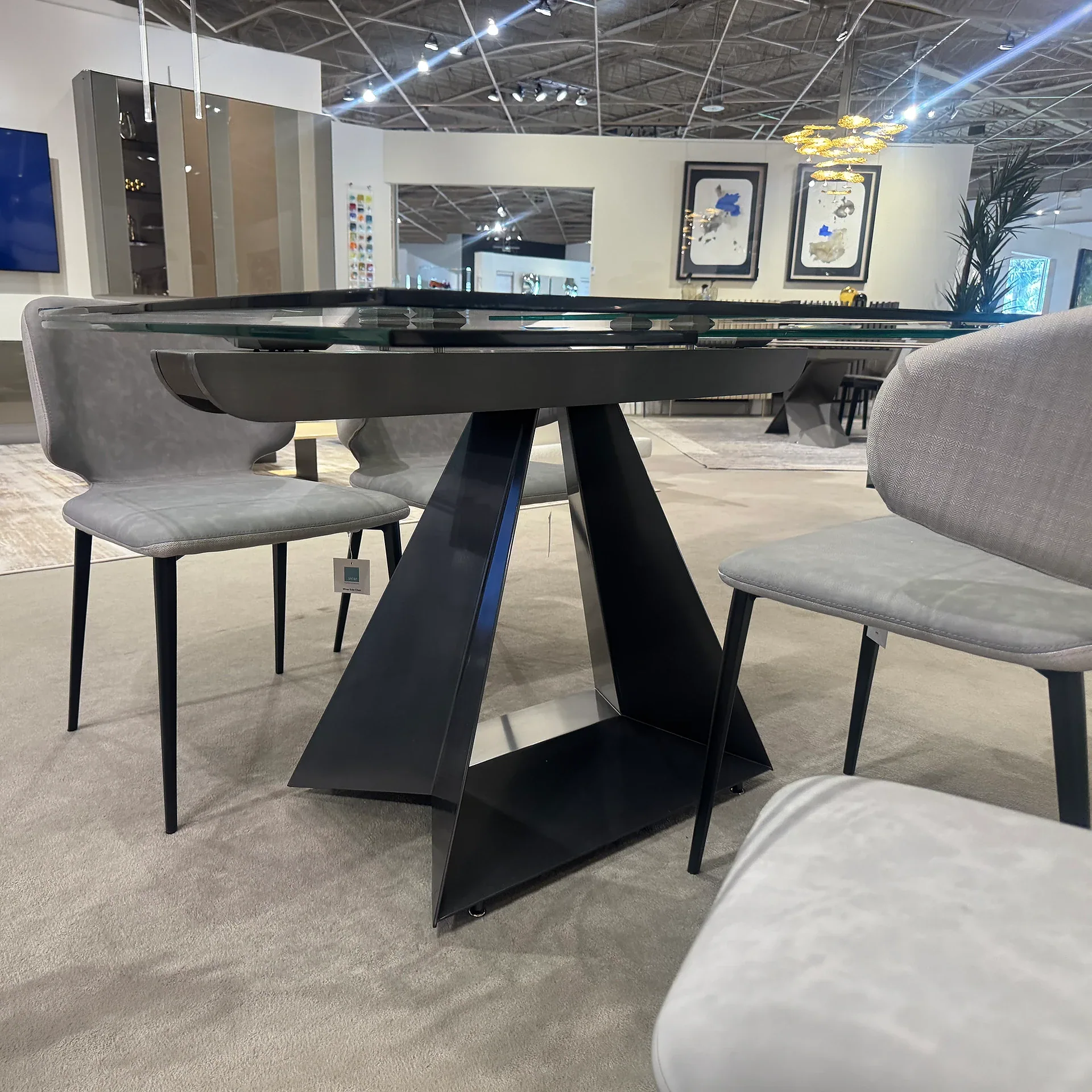 A modern dining table with a glass top and a dark geometric base, surrounded by four light gray upholstered chairs, set on a light gray carpet in a showroom.