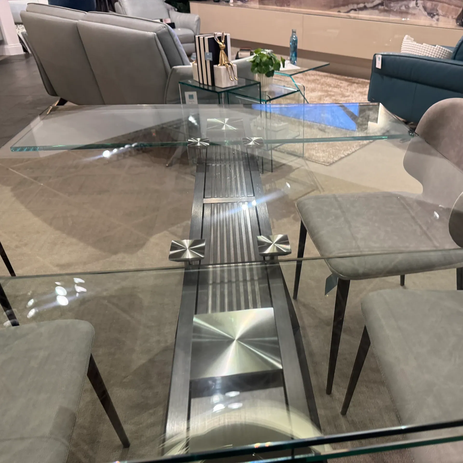 Modern dining table with a clear glass top, dark textured central support, and four light gray chairs. A living room with sofas and a glass coffee table is in the background.