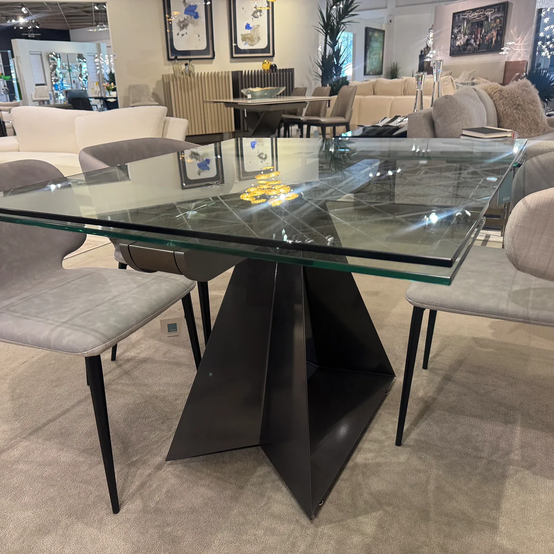 A modern dining table with a clear glass top and a dark, sculptural geometric base, surrounded by light gray upholstered chairs, displayed in a furniture showroom.