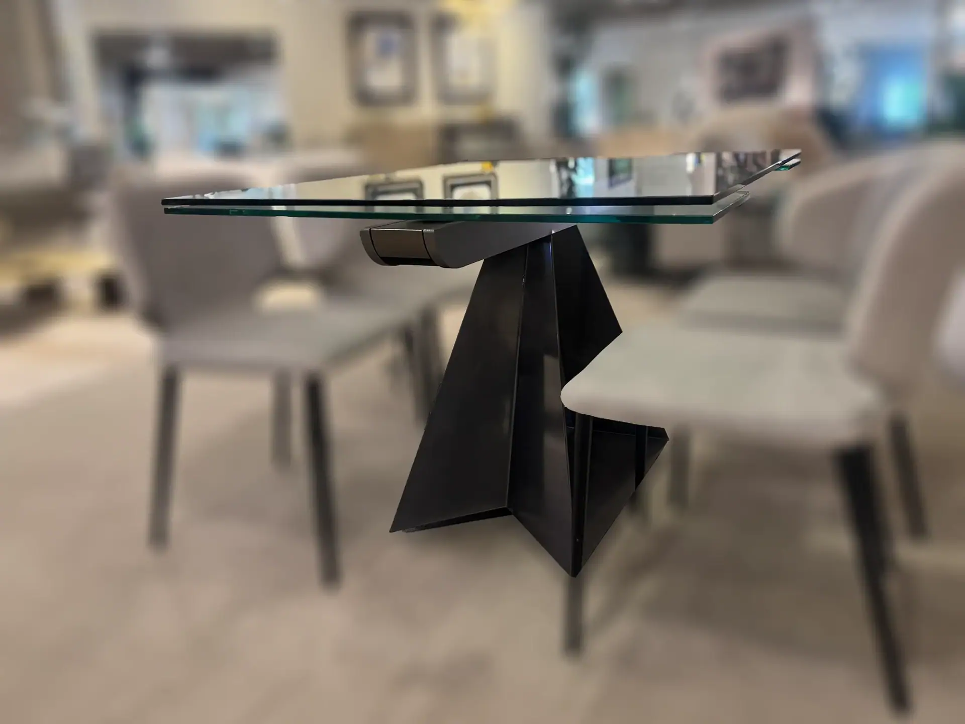 A modern dining table with a clear glass top and a black, angular, geometric base, surrounded by light-colored chairs in a blurred indoor setting.