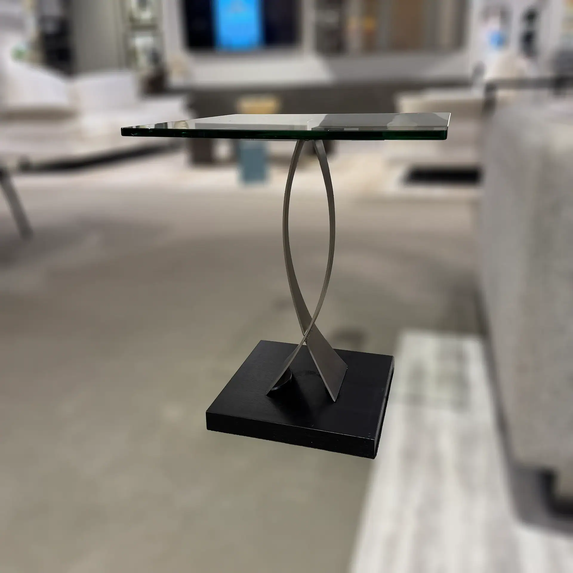 A modern side table with a clear glass top, a twisted brushed metal support, and a dark rectangular base plate. A white object rests on the glass top.