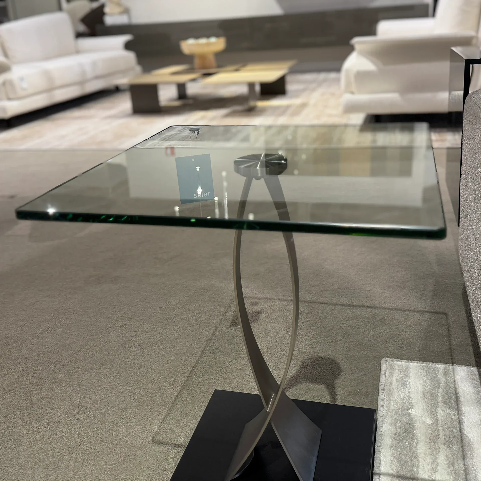 A square glass-top side table with a curved, intersecting metal base in a modern living room setting.