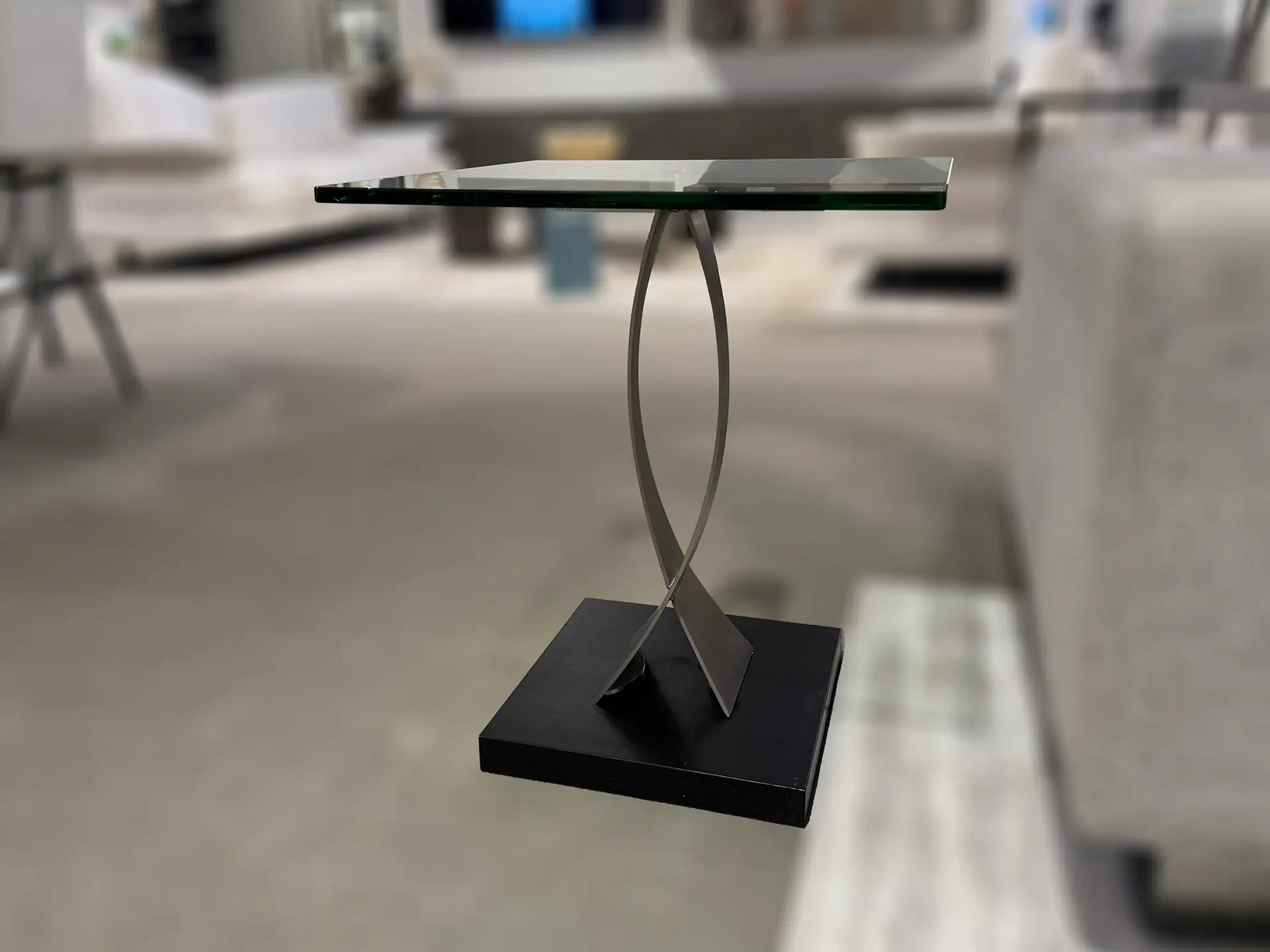A modern side table with a square glass top, a curved silver metal base, and a dark square foundation.