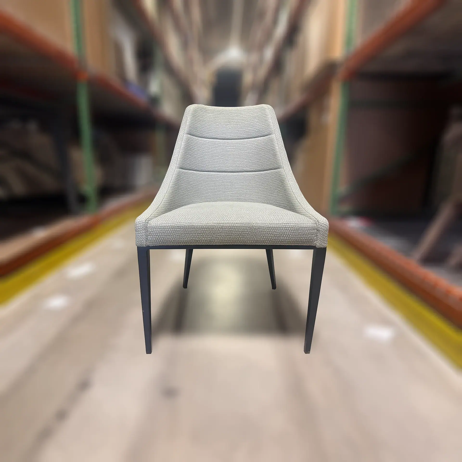 A light gray upholstered dining chair with dark tapered legs, centered against a blurred warehouse background.