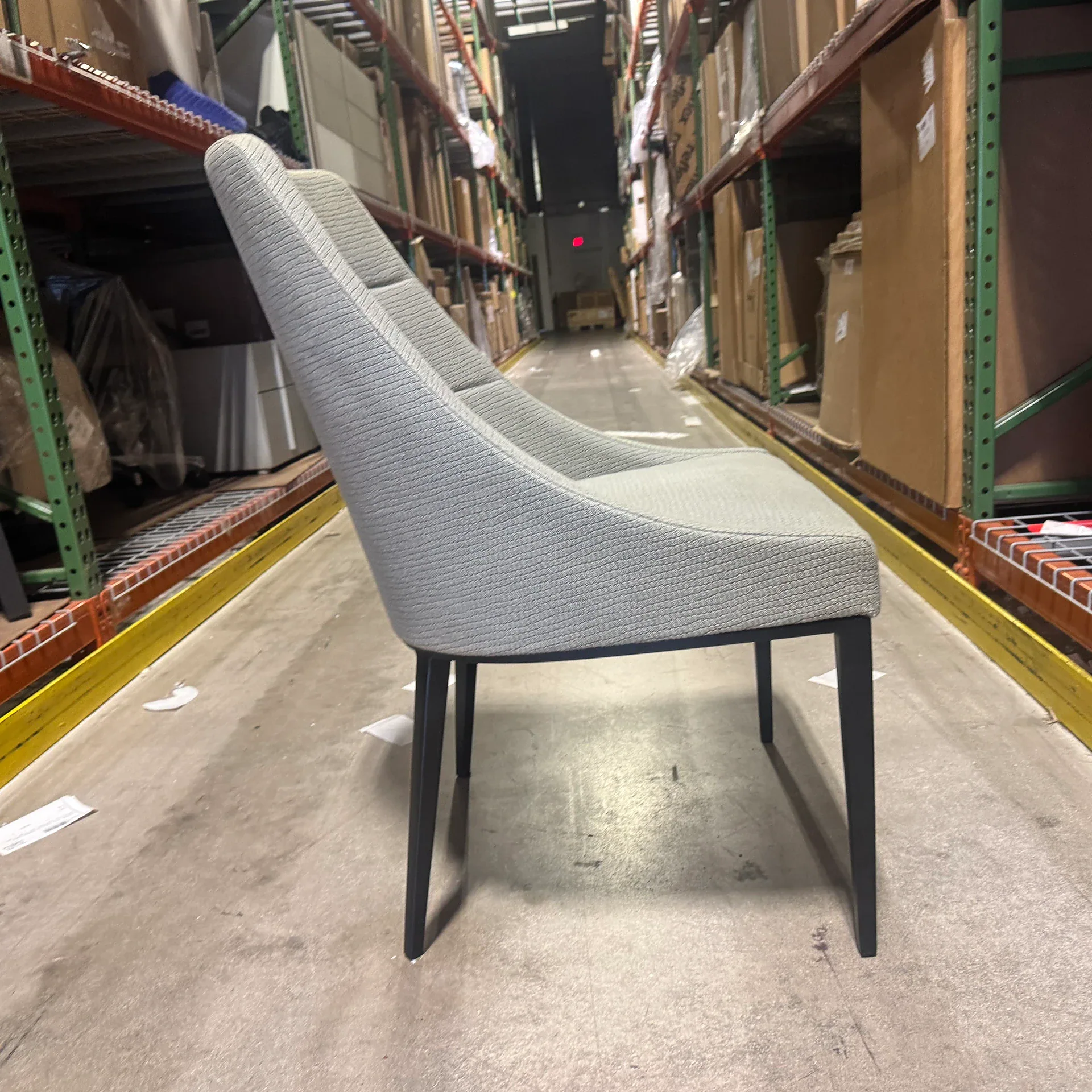 A light grey upholstered chair with black legs in a warehouse aisle between tall shelves filled with boxes.