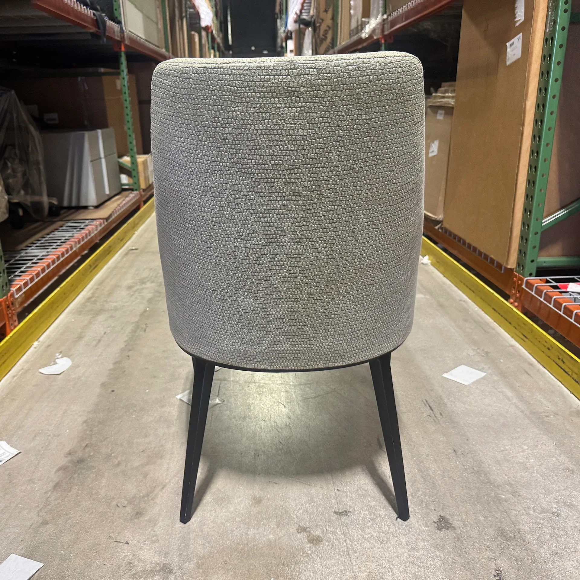 Back view of a light gray upholstered chair with dark legs on a concrete floor in a warehouse.