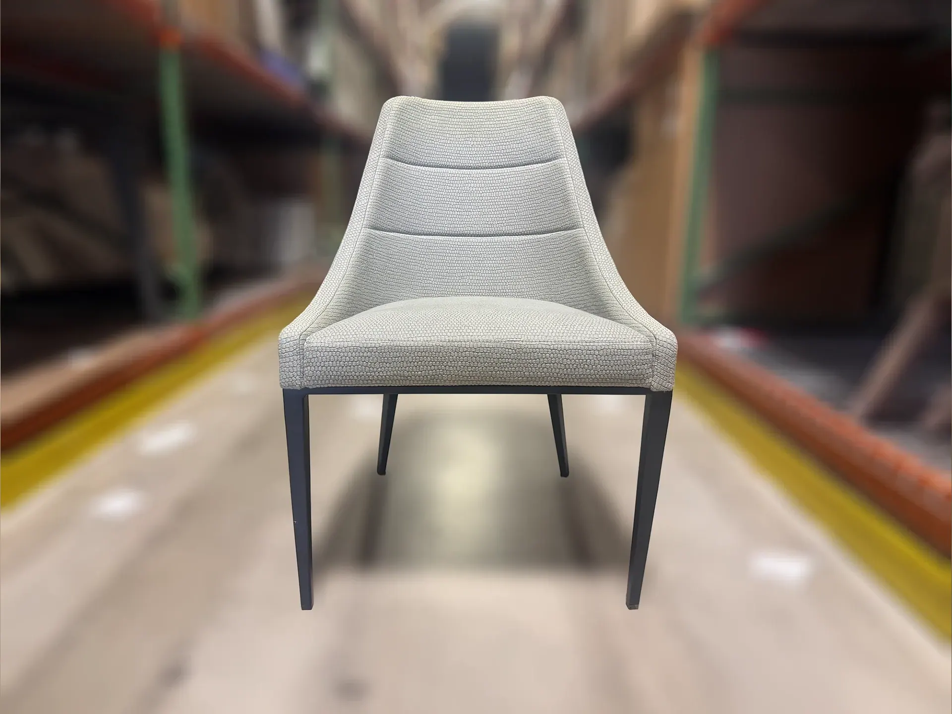 A light gray upholstered chair with dark, tapered legs sits in the foreground against a blurred warehouse background.