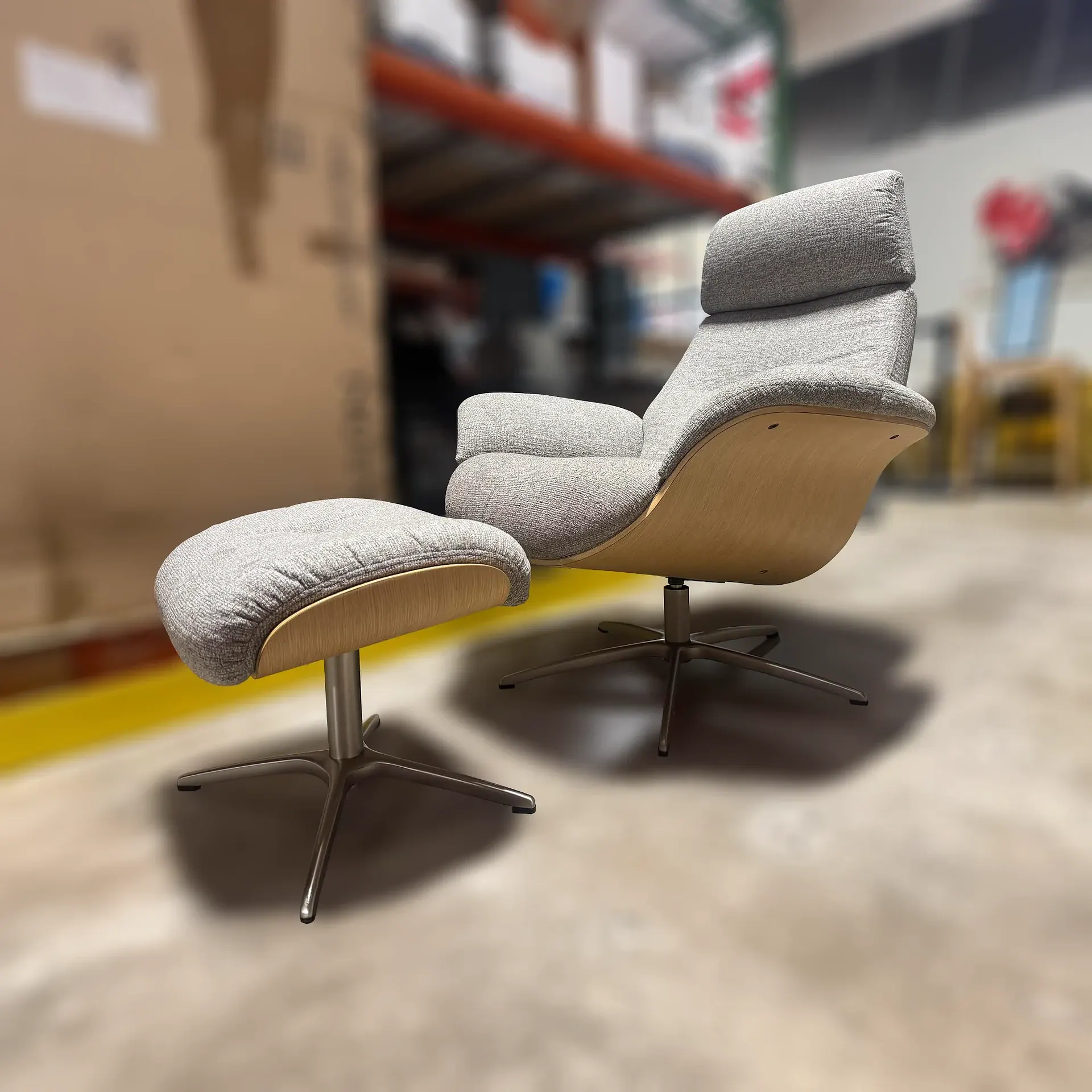 A light gray upholstered lounge chair and matching ottoman with curved light wood shells and brushed metal bases, set on a concrete floor in a blurred warehouse background.