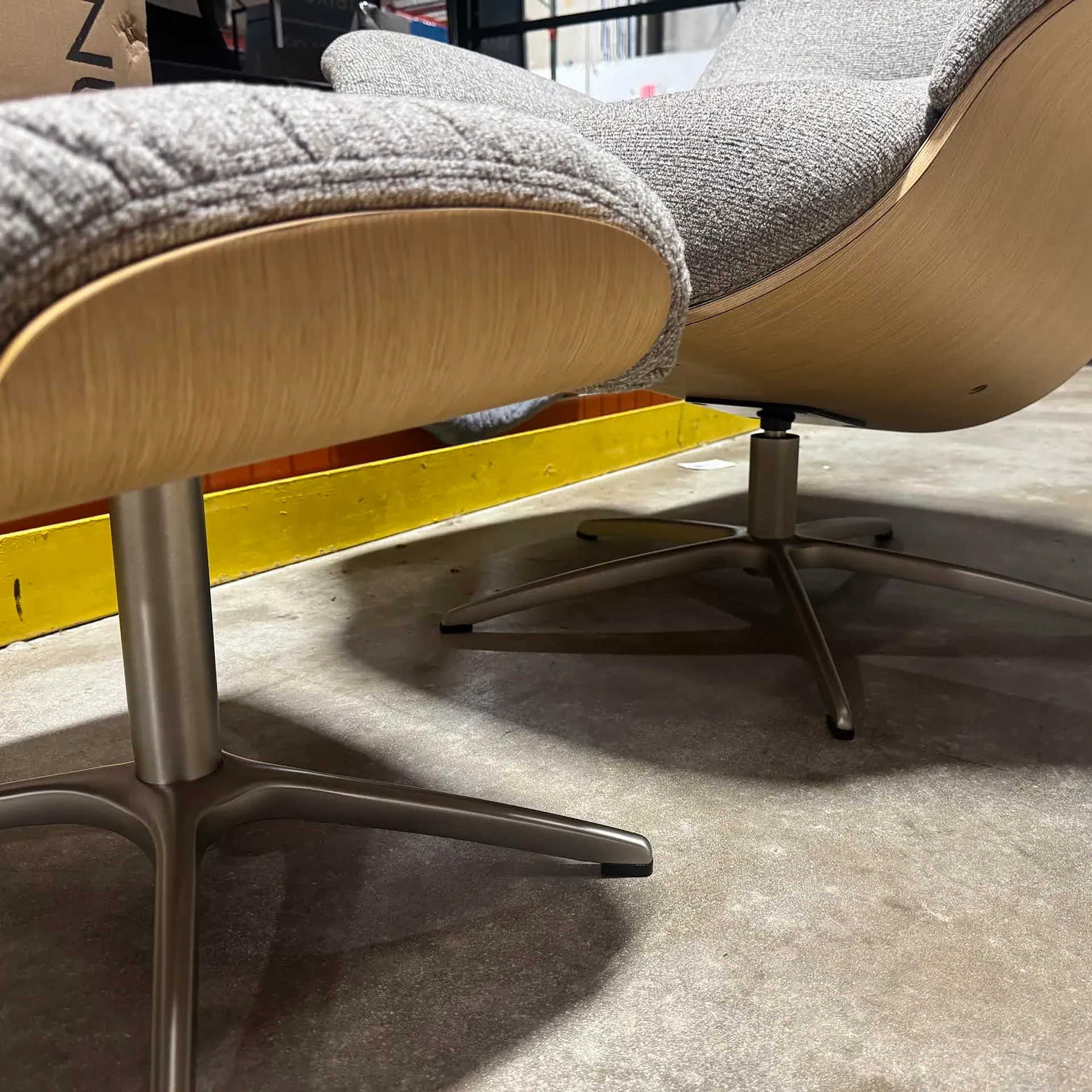 Two modern grey fabric and light wood chairs or an armchair and ottoman with brushed metal bases on a concrete floor.