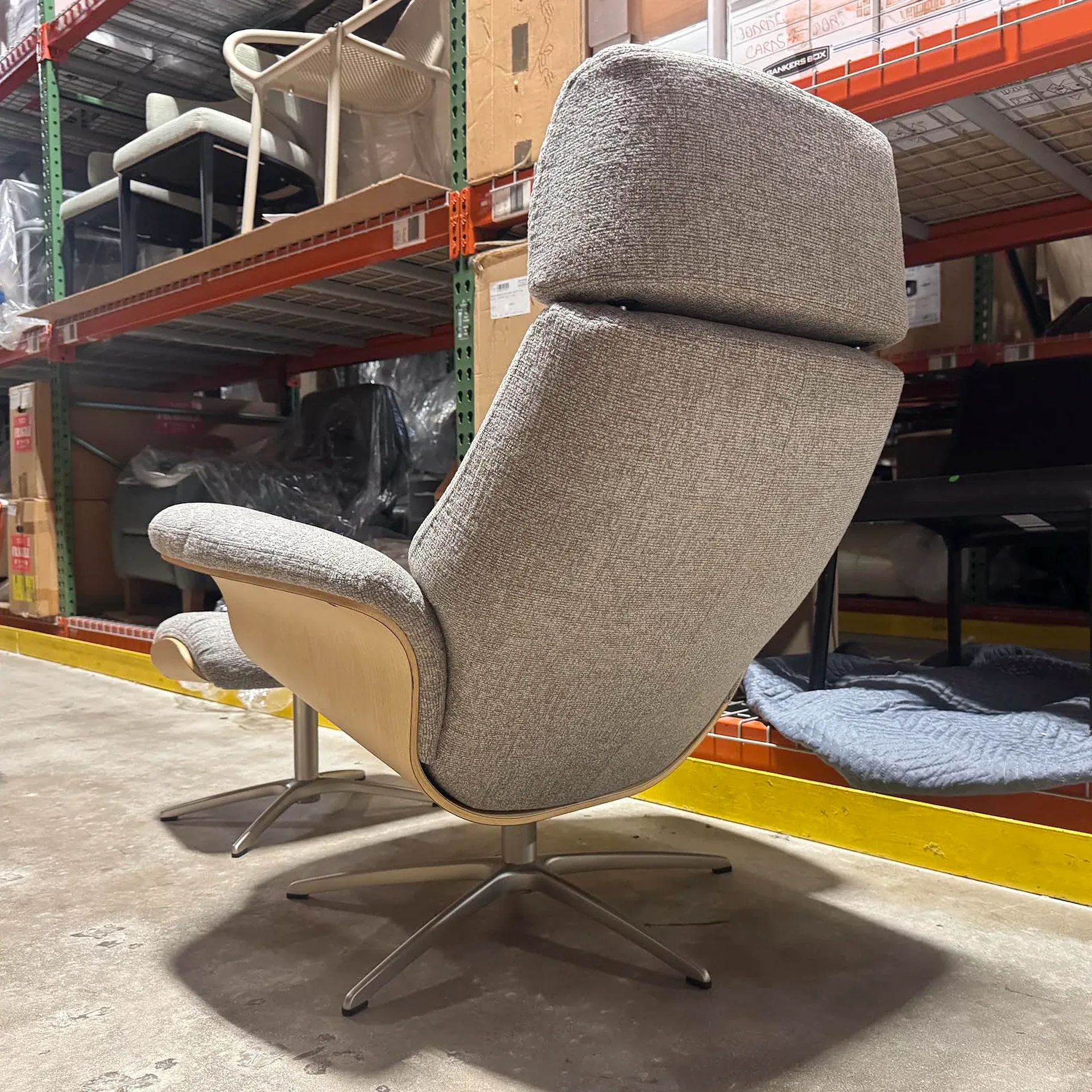 Modern grey upholstered lounge chair with a light wooden shell and a four-star metal base, positioned in a warehouse.
