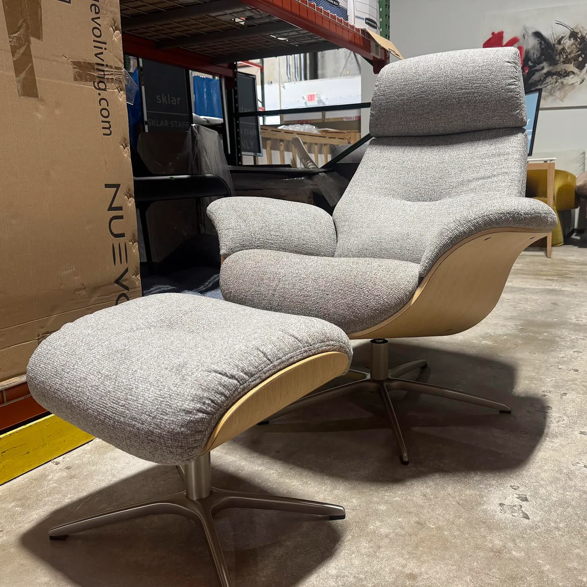 A light gray upholstered armchair and matching ottoman with curved wooden shells and metal bases, set on a concrete floor in a warehouse.